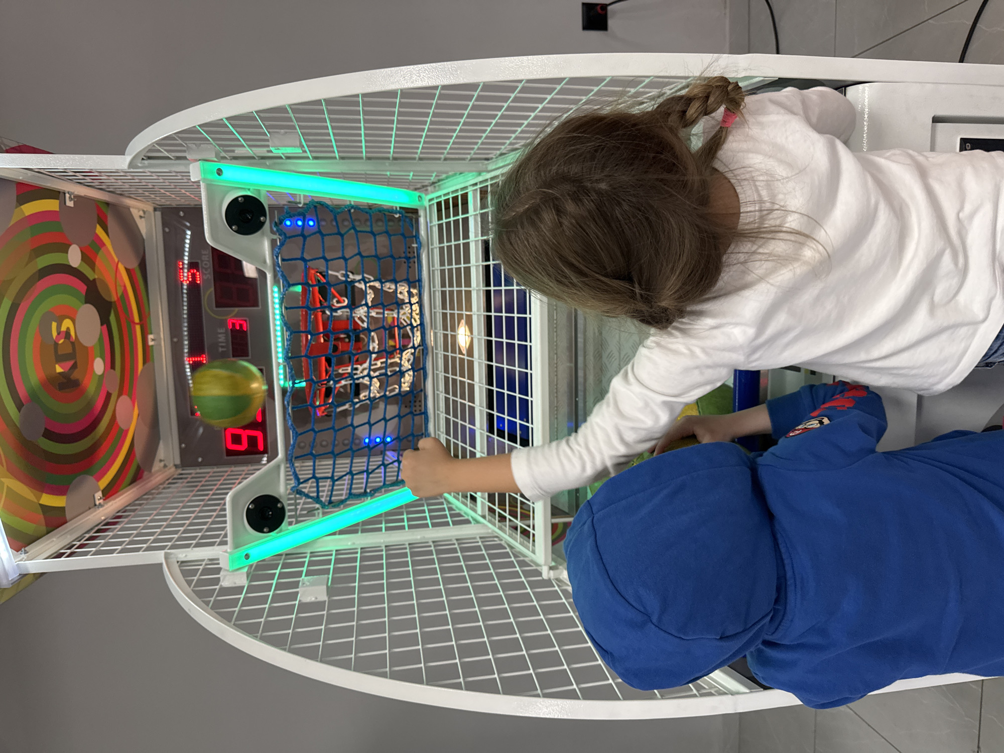 Two children are playing a basketball arcade game. One child with a white long-sleeve shirt and braided hair throws a yellow and green basketball into the hoop. Another child wearing a blue hoodie is near the game. The game's score is displayed as 3, and the time is at 6. The arcade game is inside. The location is Kranevo, Bulgaria.