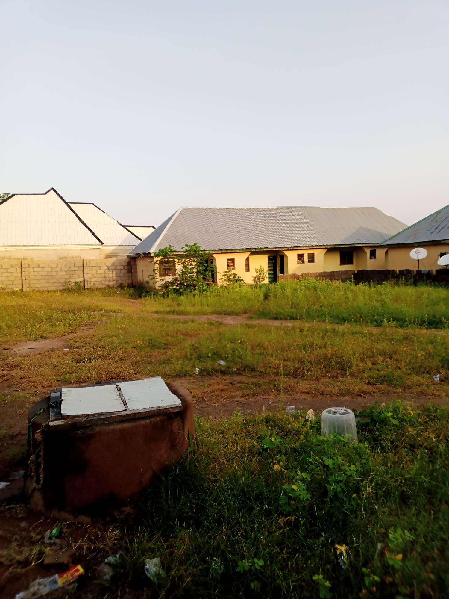 In Jauro Yinu, Nigeria, an outdoor scene depicts a low-lying building with a light yellow exterior and a corrugated metal roof, situated behind a brick wall. To the left of this building, another structure with a white, angled roof is visible. The foreground features a well with a brown, circular base and a white covering. The ground is covered with grass and dirt, with some scattered debris, including plastic bottles and wrappers. In the background, two satellite dishes are mounted on the side of the building. The sky is clear and light blue, suggesting it is either early morning or late afternoon.