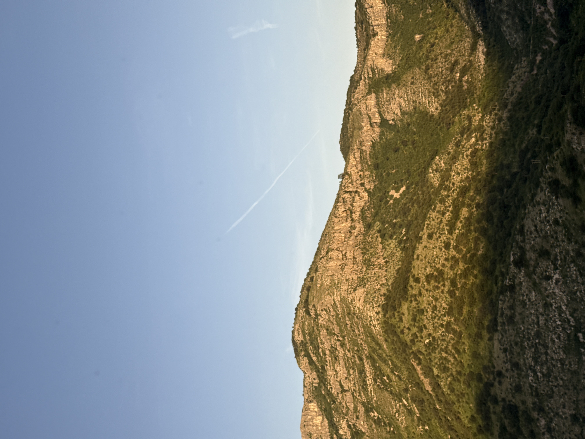 The image shows a mountain ridge in Klopot, Montenegro, seen against a clear blue sky. The mountain is characterized by layered rock formations and sparse vegetation. A contrail bisects the sky, suggesting the recent passage of an aircraft.