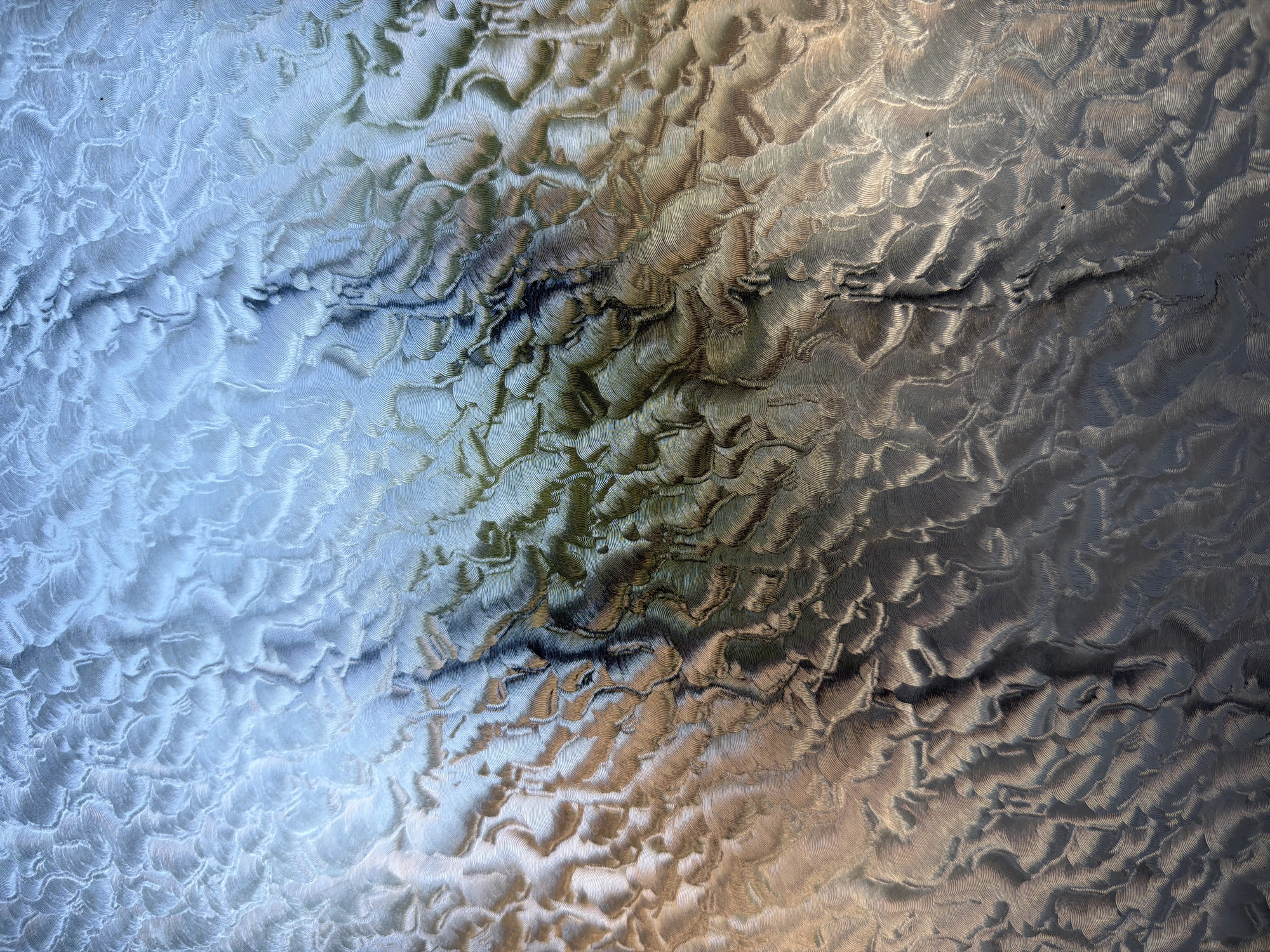 The image displays an abstract, textured surface with a gradient of colors ranging from cool blues and silvers in the upper portion to warmer browns and dark grays towards the bottom. The texture appears to be a repetitive, wave-like pattern, possibly created by machine tooling or natural erosion. Light reflects off the ridges and valleys of this pattern, creating a sense of depth and movement. There are no discernible people, animals, or specific objects identifiable within the image.
This abstract texture does not provide any visual cues to identify a specific location within the city of Piacenza, Italy, or any other geographical context.