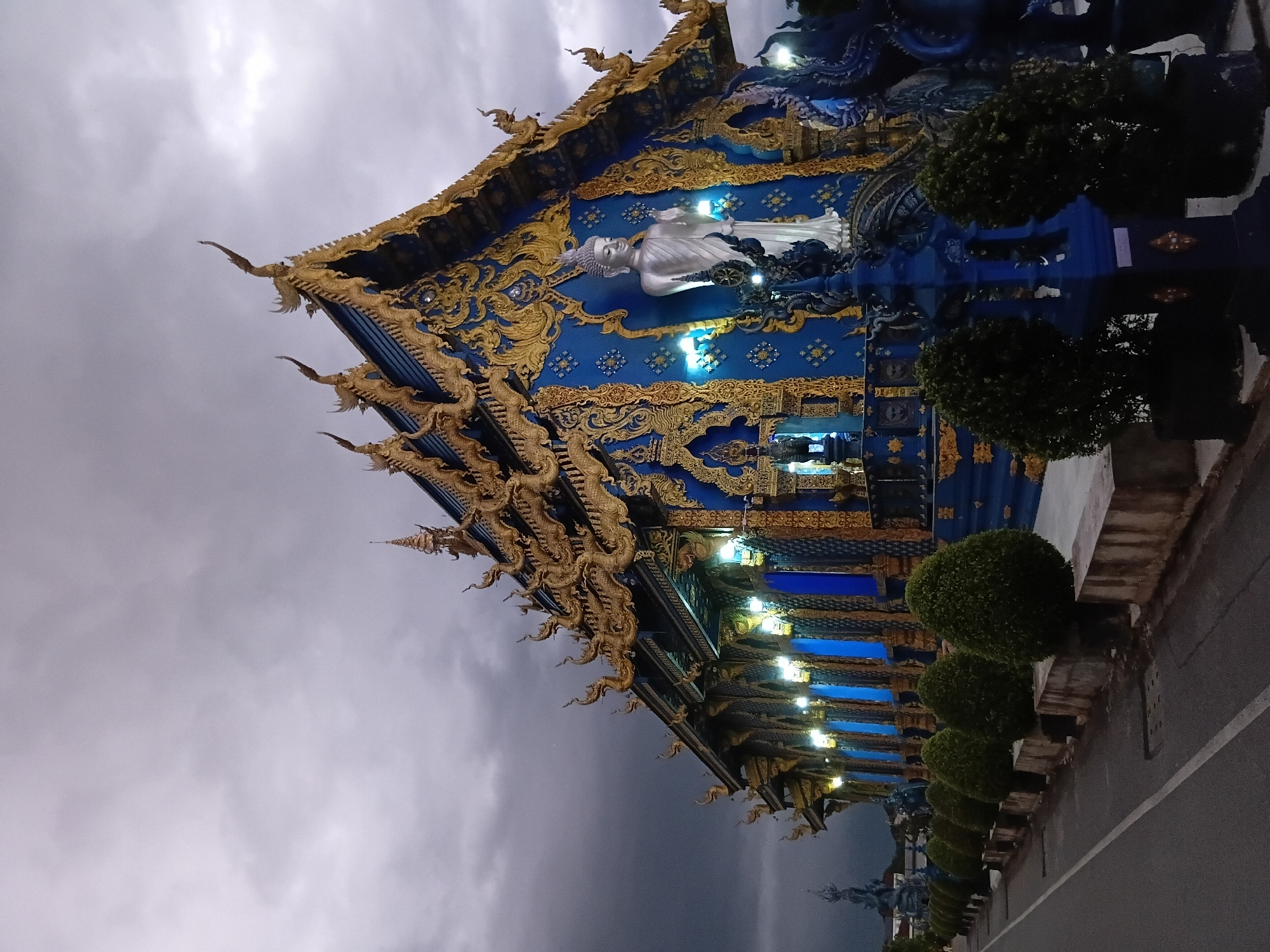 The image depicts the Wat Rong Khun, also known as the White Temple, in Chiang Rai City Municipality, Thailand, at dusk or during a storm. The temple is a strikingly blue structure adorned with elaborate golden trim and intricate designs, featuring a prominent white statue of Buddha on its facade. The roof is decorated with mythical figures and ornate golden spires. In the foreground, a paved road runs alongside a row of manicured, cone-shaped bushes planted in concrete planters. The sky is overcast and dark, with dramatic clouds suggesting impending rain or a recent storm. The lighting from within the temple casts a blue glow through its windows. No people are visible in the image.