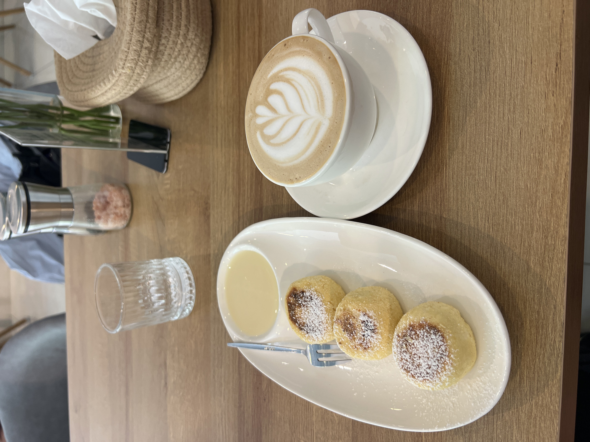 On a wooden table, likely in a café or restaurant in Bac Nha Trang Ward, Vietnam, lies a white plate containing three small, round, browned pastries dusted with powdered sugar and served with a side of light-colored sauce. A small fork rests on the plate. A white cup of latte, adorned with latte art, sits on a white saucer. A small, empty glass and a glass container of pink salt are also present on the table. In the background, a woven basket or holder is visible, likely containing napkins. The scene suggests a meal or snack consisting of coffee and pastries.