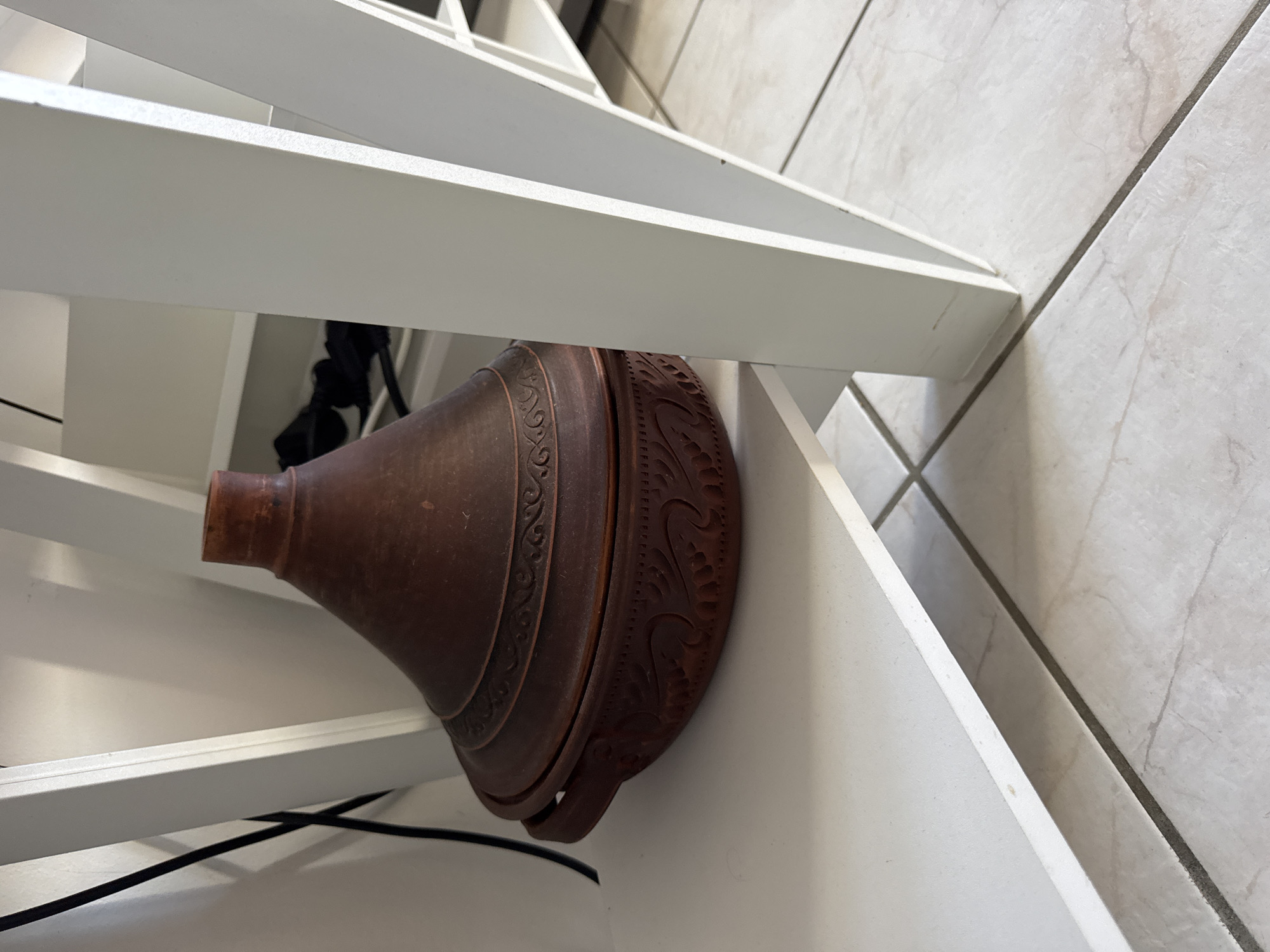 A brown clay tagine is placed on a white shelf. The tagine has a conical lid with decorative bands and a rounded base with carved patterns. The shelf is part of a white shelving unit, visible on the right side of the image. A black cable runs down the white wall behind the shelving unit on the left. The flooring appears to be light-colored tiles with grout lines. The scene is indoors, likely in a residential setting in Höhenkirchen-Siegertsbrunn, Germany, given the context of the prompt, though no specific identifying landmarks are visible in the image. There are no people present.