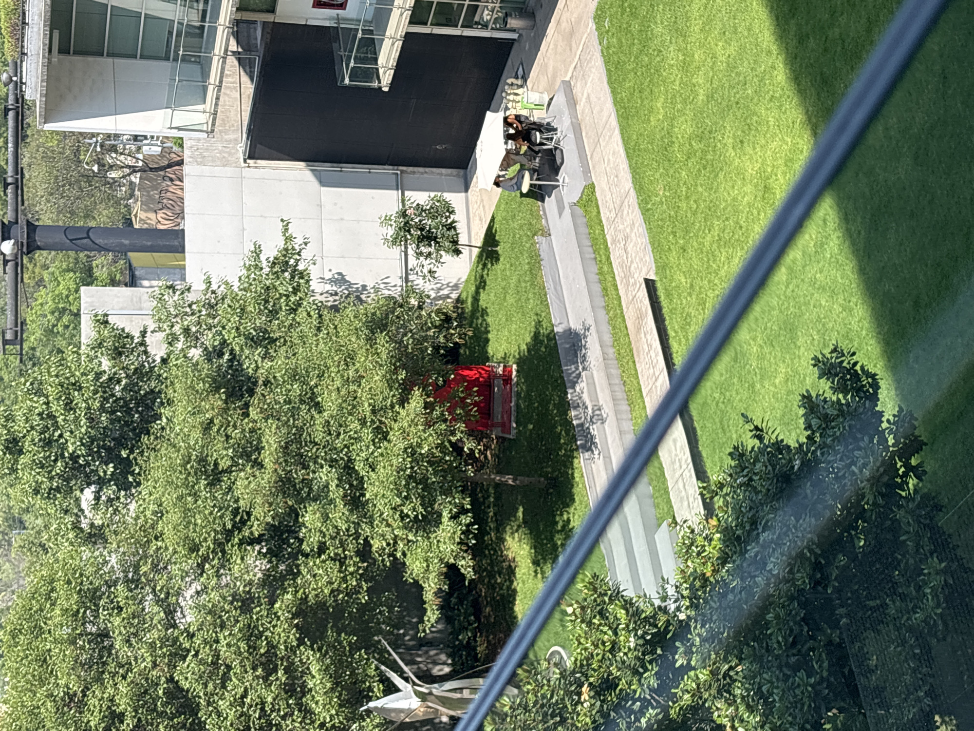This is an aerial view taken from a high vantage point in Mexico City, Mexico. The scene shows a mix of architectural elements and greenery. A modern building with glass windows and a black overhang is visible. Adjacent to the building, there is a paved area with people sitting at tables with an umbrella. A grassy lawn extends from the paved area, bordered by a walkway. Trees and foliage cover a portion of the foreground, with a red structure noticeable among the trees. A reflective sculpture is present in the lower left. A diagonal line bisects the image, likely a structural element of the observation point.