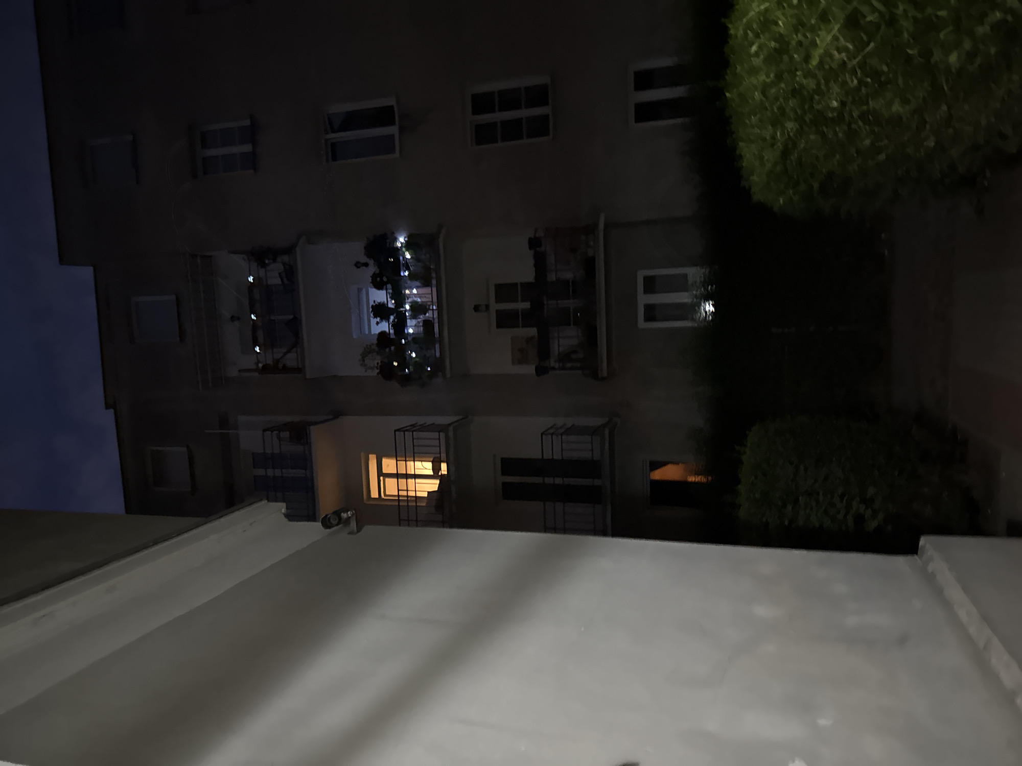 This is a nighttime exterior shot of an apartment building in Unknown, Unknown. The building appears to be made of concrete or stucco and has multiple stories with balconies and windows. The sky is dark, indicating it is night. A large, light-colored wall of another building or structure occupies the left side of the frame, casting a shadow. The building in the background has several balconies with railings. One balcony on the second floor from the bottom is lit from within and appears to have furniture. Another balcony above it is decorated with string lights and plants. Several windows are visible on the building, some of which are illuminated. There are bushes in front of the building. A security camera is visible mounted on the lighter-colored structure on the left.