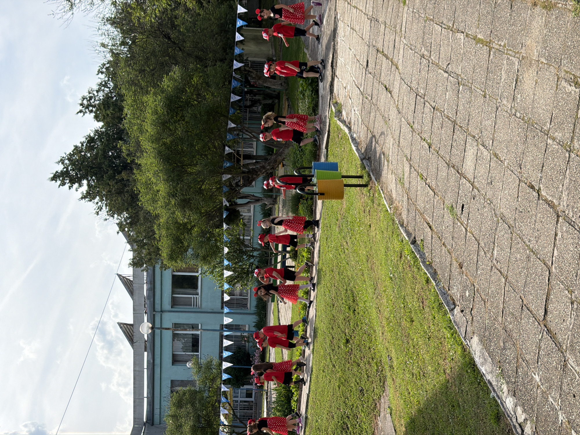 The image captures an outdoor scene featuring a group of approximately ten children dressed in matching red and white polka-dot outfits. Most of the children, predominantly girls, wear red dresses with white polka dots, while the boys wear red t-shirts and black shorts. Many also wear headbands with ears, reminiscent of Minnie or Mickey Mouse.
The children are arranged in a line along a paved pathway, likely performing or participating in an organized event such as a parade or school festival. They are generally facing towards the right side of the frame, with some looking towards the camera or to the left. To the left of the pathway, there is a vibrant green grassy area.
In the mid-ground, next to the grassy area, are several colorful cylindrical objects in blue, yellow, and green, possibly acting as bins or props. Behind the children and the grass, a dense line of green trees provides a natural backdrop. Further in the background, a light blue building with multiple windows and a distinctive roofline is visible, suggesting a school or institutional setting. White and blue triangular flags, strung as bunting, are draped across the scene, indicating a festive occasion.
The scene is illuminated by bright daylight, suggesting it is either morning or afternoon. The sky above is light with scattered white clouds. The paved pathway shows signs of age, with grass visible growing through cracks. No visible text is present in the image.
