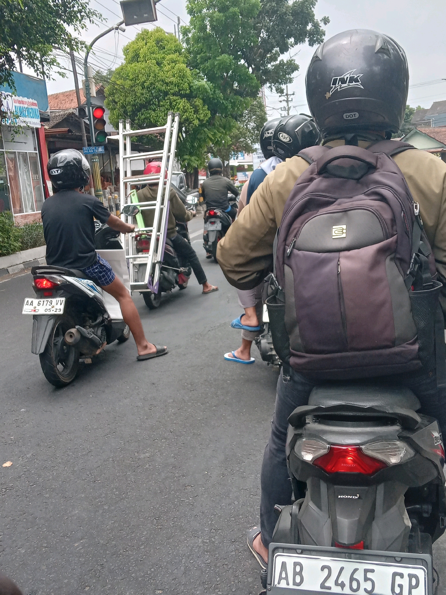 The image captures a street scene in Yogyakarta, Indonesia, likely on a cloudy day. A group of motorcyclists are navigating through traffic. The foreground features a person wearing a black helmet and a tan jacket, carrying a dark backpack, riding a motorcycle. Behind them, another motorcyclist with a black helmet is visible. Further ahead, two more motorcyclists are seen, one carrying a long ladder strapped to his motorcycle. The street is paved with asphalt, and various buildings and trees line the sides, suggesting an urban or semi-urban environment. A traffic light is visible in the background, with the red light illuminated. The license plates of some motorcycles are partially visible, indicating Indonesian registration. The overall impression is a typical street scene with daily commuter activities.