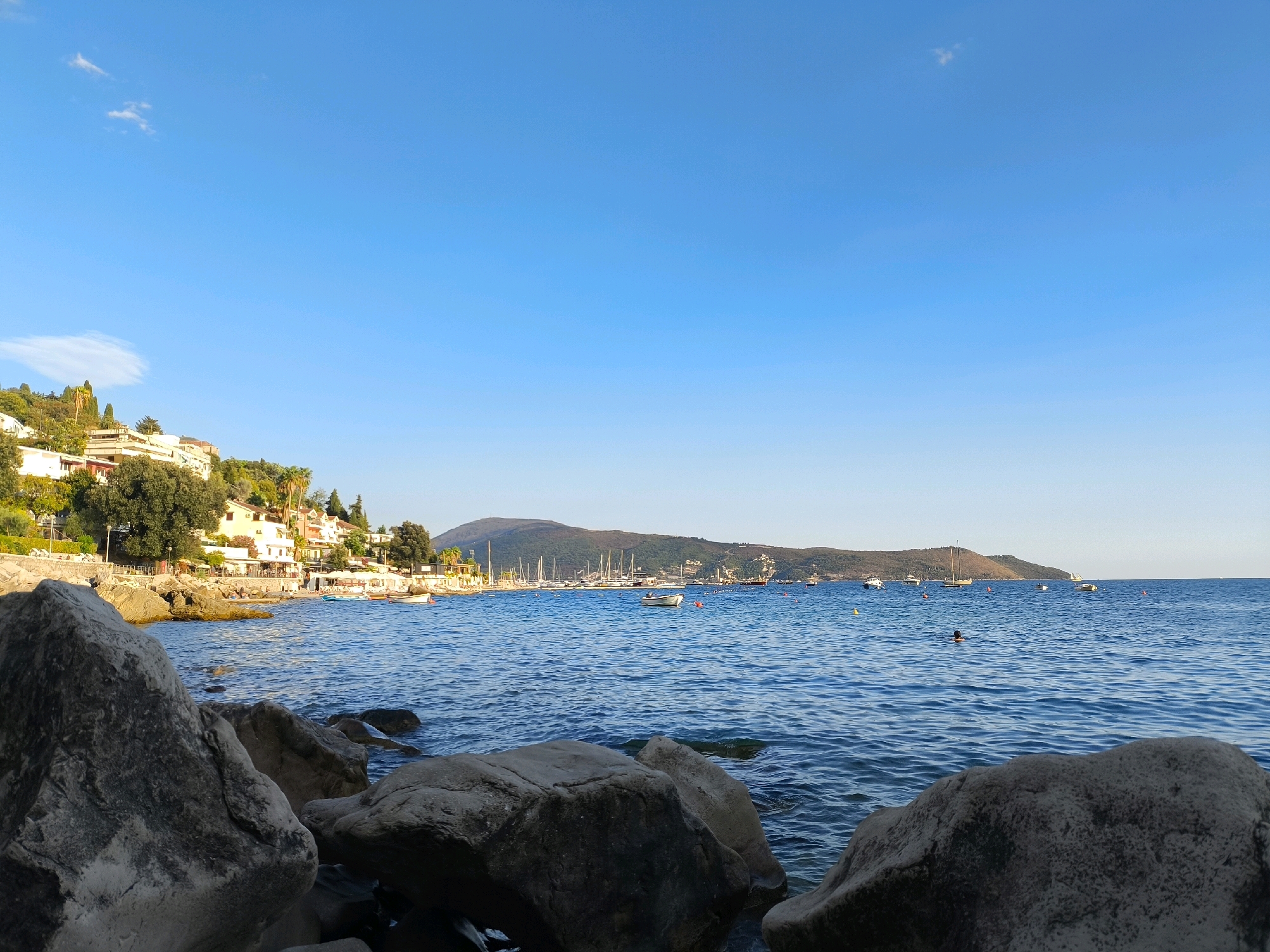 The image depicts a scenic view of the Adriatic Sea in Herceg Novi, Montenegro, likely from a rocky shoreline. In the foreground, large, rough-textured boulders are prominent, with their weathered surfaces catching the sunlight. The mid-ground reveals a calm, blue sea with gentle ripples, dotted with several small boats and buoys. Further in the distance, a picturesque coastline is visible, characterized by buildings nestled on a hillside adorned with lush green trees and vegetation. Some of these buildings appear to be houses and establishments with outdoor seating, suggesting a coastal town or resort area. The background features a majestic mountain range, bathed in the warm glow of the setting sun, with sailboats anchored in the bay. A lone swimmer is visible in the water, adding a touch of human activity to the tranquil maritime landscape. The sky above is a clear, vibrant blue, with a few wispy clouds adding depth to the expanse.