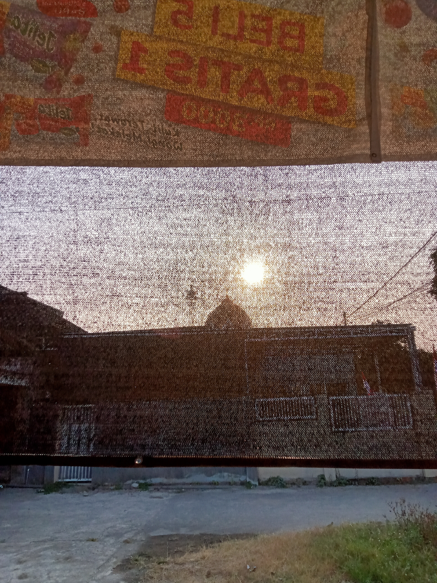 This is a photograph taken in Simo, Indonesia, looking through a dark mesh screen. In the upper portion of the image, a colorful banner with Indonesian text is visible, featuring words like "BELI" (buy) and "GRATIS" (free) in large red and yellow letters. The lower portion of the image shows a silhouette of buildings against a bright sunset sky, with the sun positioned just above the roofline. The mesh screen creates a textured overlay, obscuring some details of the buildings and landscape. The foreground shows a dirt or gravel area with some sparse vegetation. The overall impression is that of a view through a protective barrier, possibly a shade cloth or netting, at dusk.