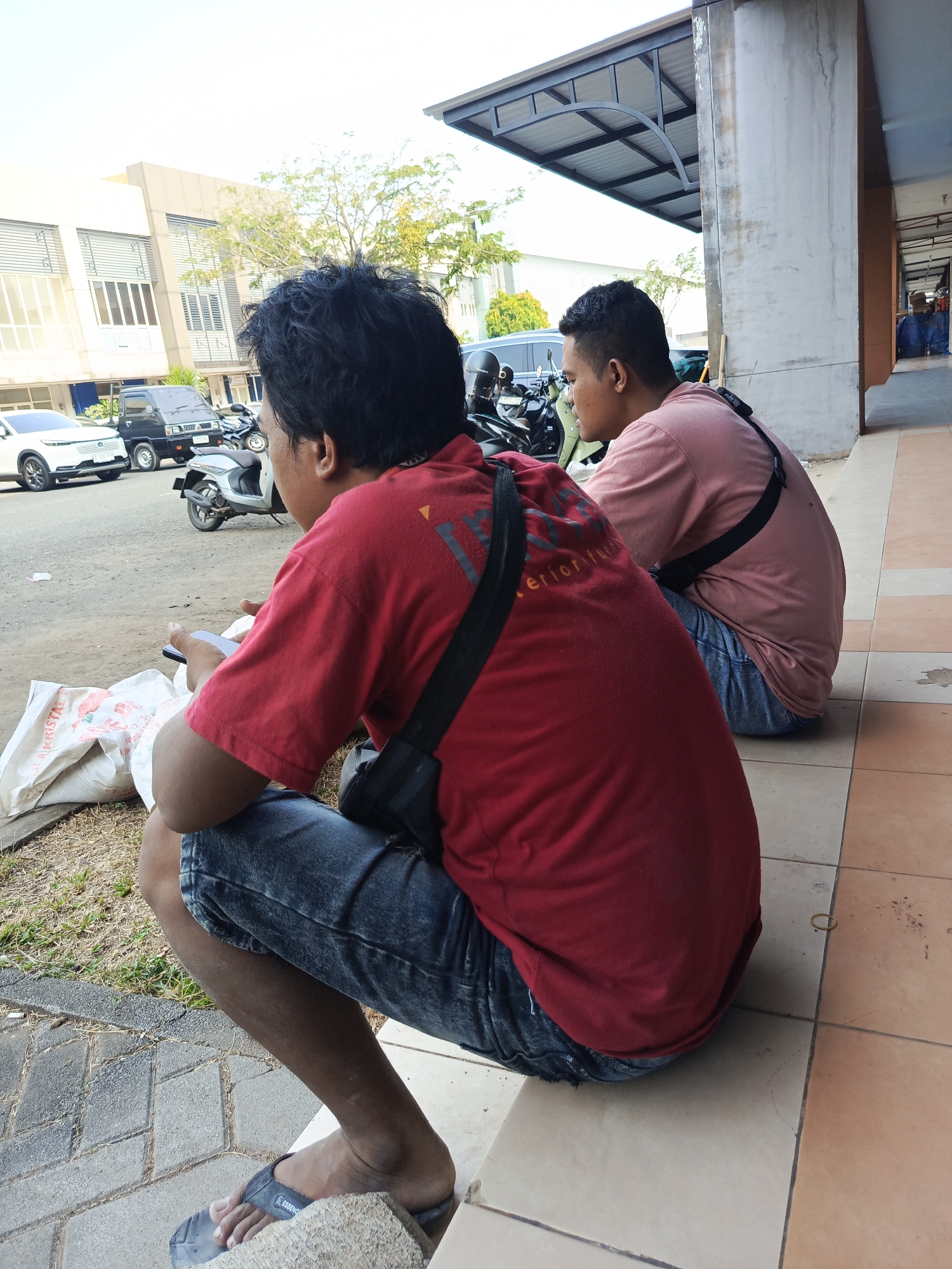 Two men are squatting on a tiled walkway in front of a building. The man in the foreground is wearing a red t-shirt and blue jeans. He is holding a smartphone in his left hand. The man in the background is wearing a pink t-shirt and blue jeans. Both men have dark hair and are facing away from the camera. In the background, there are several parked vehicles, including a white car and a black truck, as well as motorcycles. A modern building with glass windows and louvers is visible. The environment appears to be an urban setting, possibly a parking area or street in Tangerang, Indonesia. The ground is a mixture of concrete tiles and gravel.