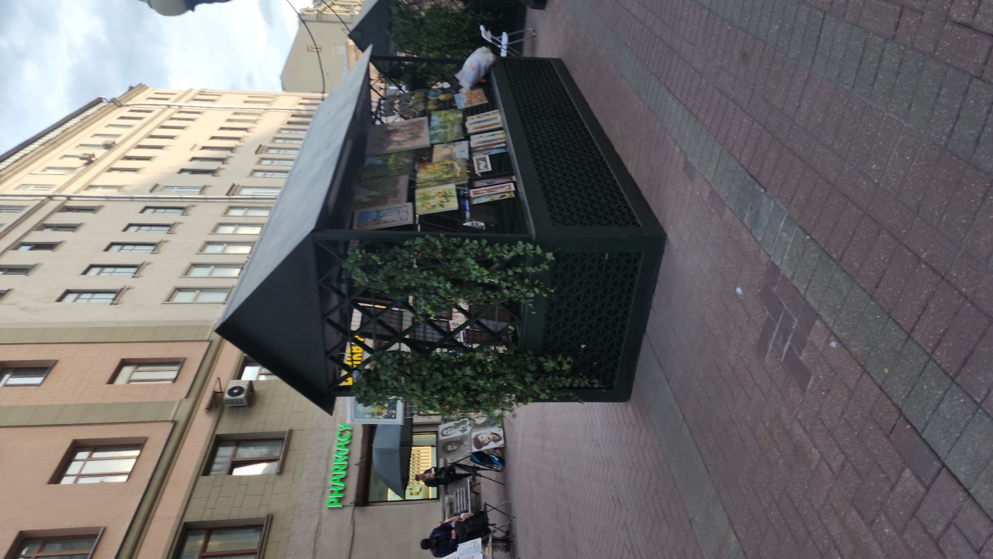 This media file depicts an outdoor scene in Moscow, Russia. The primary focus is a small, dark green, open-air kiosk displaying paintings. The kiosk is adorned with greenery, likely artificial plants, and the paintings are arranged on its exterior walls. The paintings themselves appear to be diverse, featuring landscapes and portraits.
In front of the kiosk, on the paved sidewalk, two individuals are seated on chairs. One is a person in a dark jacket looking down at something in their lap, possibly a book or a phone. The other person, partially obscured, is also seated. A black umbrella is open nearby, suggesting it might be raining or the sun is very strong.
Behind the kiosk, a building with a pale beige facade and numerous windows is visible. To the left of the kiosk, another building is partially visible with a peach-colored facade and windows. A sign with bright green lettering reading "PHARMACY" is prominently displayed on the side of this building. An air conditioning unit is mounted on the facade of the peach-colored building.
The ground is paved with reddish-brown bricks and a strip of grey bricks. The sky is overcast, indicating a cloudy day. The overall impression is of a street scene with an art display and people engaged in a moment of rest or observation, possibly a marketplace or a public square.