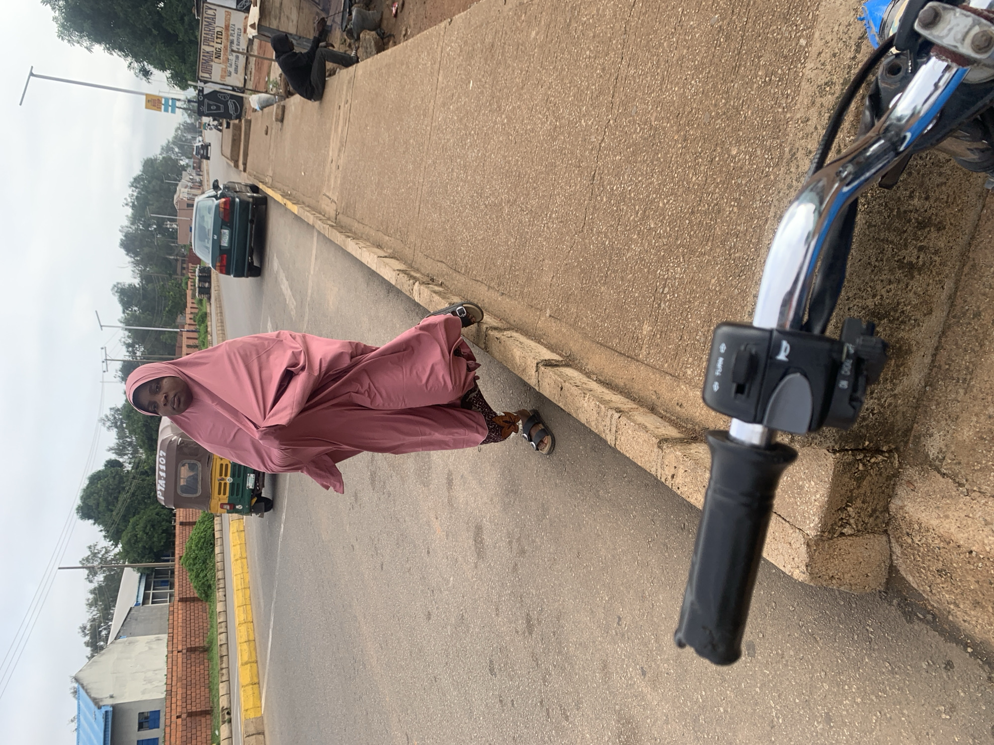 This outdoor image, taken in Barakin Lerawi, Nigeria, depicts a street scene under an overcast sky during daylight. In the immediate foreground on the right, the chrome handlebar of a motorcycle is visible, featuring a black handgrip and control switches labeled "TURN" and "CHOKE."
The central subject is a person standing by the roadside, dressed in a long, loose-fitting pink garment and a matching head covering (likely a hijab or abaya). They are wearing sandals, revealing leopard-print socks. Their face is partially visible, looking towards the camera.
The paved road features yellow-painted curbs, bordering a concrete sidewalk. Several vehicles are present: a dark green sedan is seen further down the road, and a yellow and green tricycle, commonly known as a keke napep, is visible behind the person, bearing the text "PTA-1107."
Along the sidewalk, various structures and activities are apparent. A prominent sign for "LOHMAK PHARMACY NIG. LTD." is visible, alongside other text such as "NAF GATE," "SHIPPING PLAZA," "ABRATTOR," and "& RAYFIELD." Another individual is seen sitting or crouching on the raised sidewalk further down. The background features a mix of buildings, utility poles, and scattered trees, contributing to the urban or semi-urban ambiance of the scene.