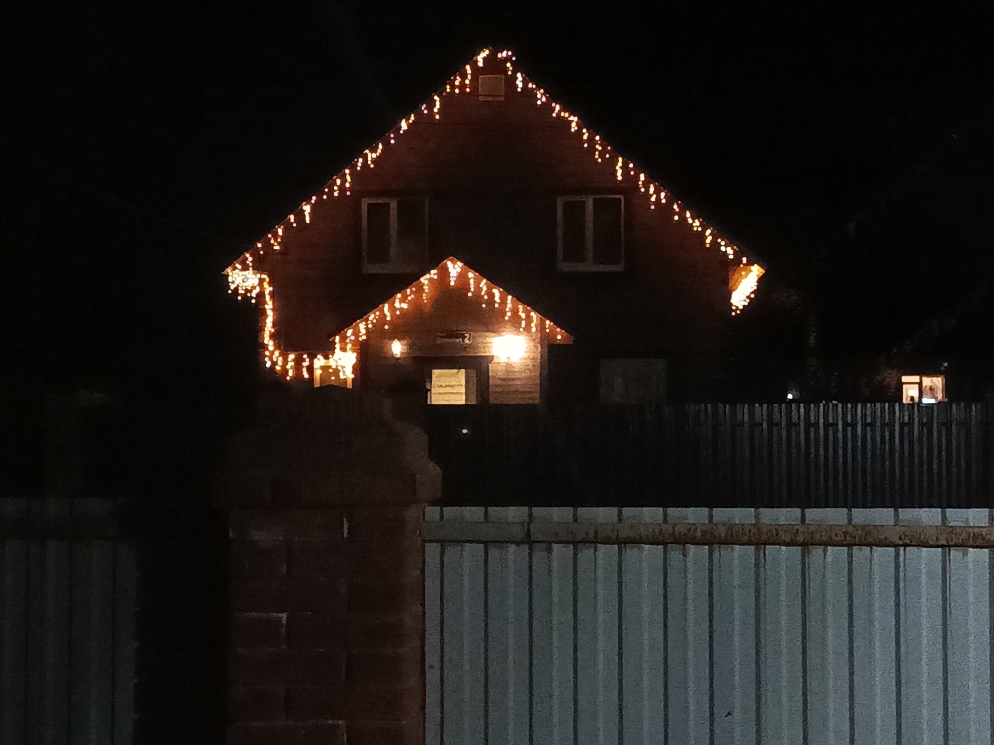 At night, a two-story house decorated with orange string lights is viewed behind a fence. The house has a gable roof and two windows on the second story. The string lights follow the outline of the roof. Light is visible from the ground floor of the house. The fence is constructed from corrugated metal, with a brick support post on the left. The location is Unknown, Unknown.