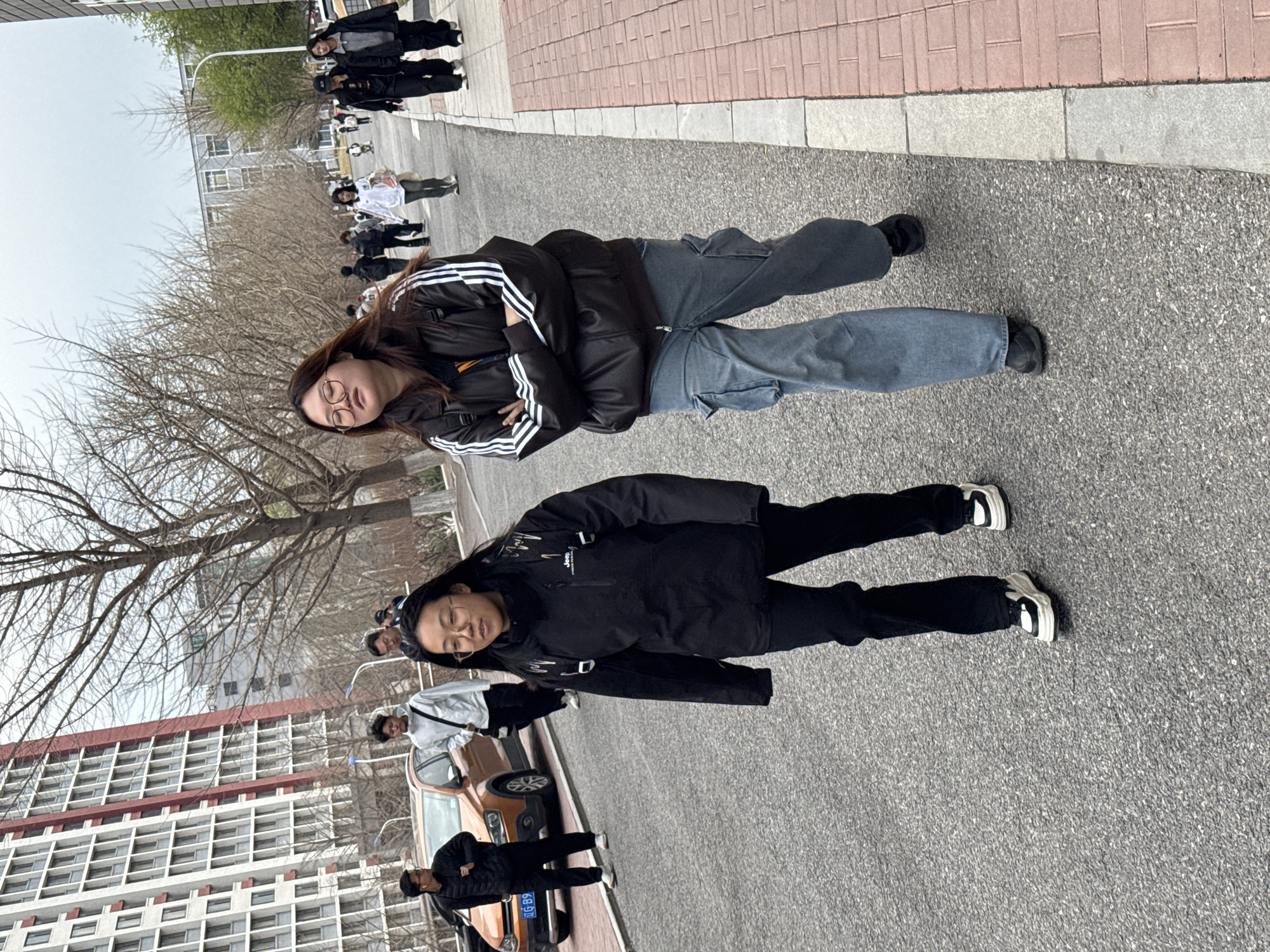 The image shows two young women standing on a paved road. The woman on the left wears a black jacket with white stripes on the sleeves and light blue jeans. She has long dark hair and wears glasses. The woman on the right is wearing a black jacket, black pants, and white shoes. In the background, there are other people walking on the road. A light brown car is parked near the sidewalk. Behind them, there are buildings and trees. The location appears to be an urban street in Zhongtun, China.