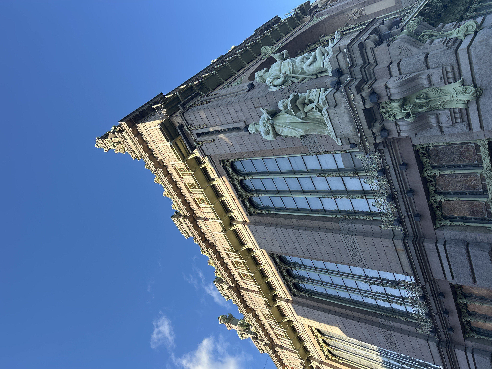 An upward-angle, eye-level, medium shot captures the corner of a grand, ornate building with detailed architecture under a clear, bright blue sky with a few wisps of white clouds. The building is constructed of pinkish-brown stone blocks and features elaborate green metalwork, including intricate balconies, window frames, and decorative elements. Two large, pale green statues stand prominently at the corner, one depicting a woman with a book and a child reading on a stack of books, and the other a man with a staff and a crouching child. The statues appear to be part of the building's facade. The building's windows are tall and segmented with green mullions, reflecting the sky. The overall impression is one of historical significance and artistic grandeur. The location is identified as Unknown, Unknown.