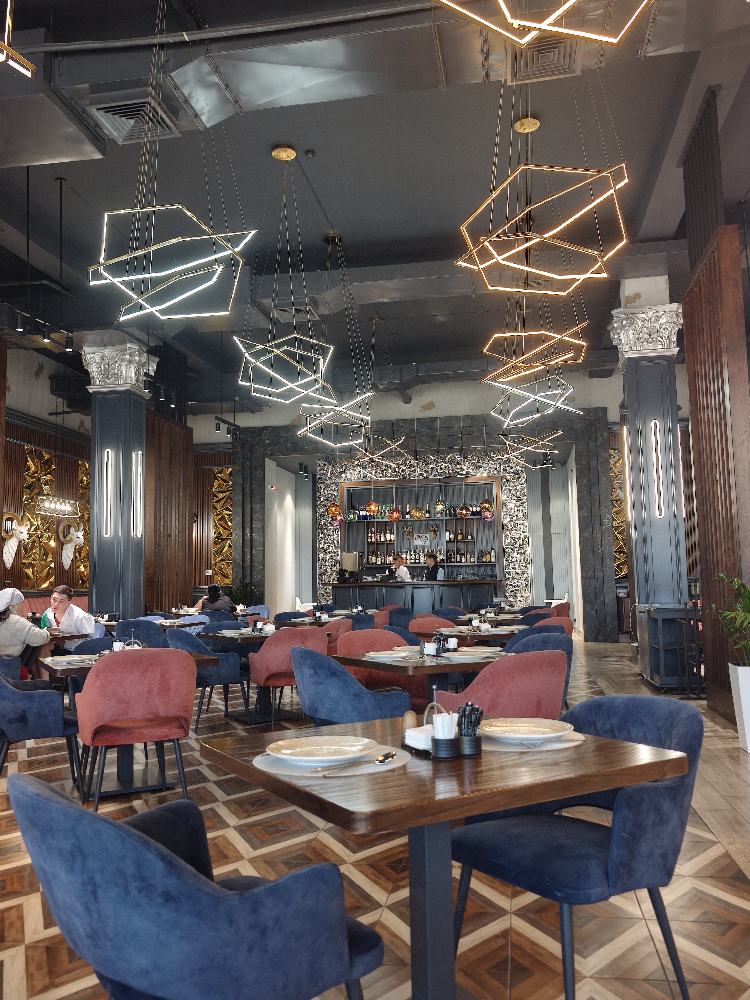 The image shows the interior of a restaurant in Çolpon-Ata, Kyrgyzstan. The restaurant features a high ceiling with geometric chandeliers. The seating area includes dark wooden tables surrounded by blue and red velvet armchairs. The floor is decorated with a patterned wooden parquet. In the background, there is a bar area with shelves stocked with bottles. Several patrons are seated at various tables. The restaurant's décor incorporates decorative columns, textured wall panels, and metal artwork, providing an upscale and modern ambiance.