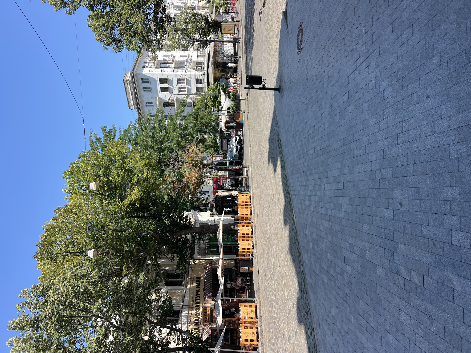The image depicts a street scene in Odessa, Ukraine, on a clear, sunny day. The foreground is dominated by a wide pedestrian area paved with light-colored stone blocks, casting distinct shadows from unseen objects. To the left, a row of outdoor seating for a cafe is set up along the sidewalk. Wooden barriers with a coffee cup motif delineate the seating areas, and white umbrellas provide shade. Several tables are occupied by patrons.
Behind the cafe, buildings with architectural details suggestive of European city design are visible. The main building in the mid-ground is a white, multi-story structure with balconies. Trees with lush green foliage line the street, interspersed with lampposts and overhead wires.
In the background, a kiosk with a colorful mural is situated, and people are walking in the vicinity. Further down the street, more trees and buildings contribute to the urban landscape. The overall impression is of a lively European city square during daytime.
