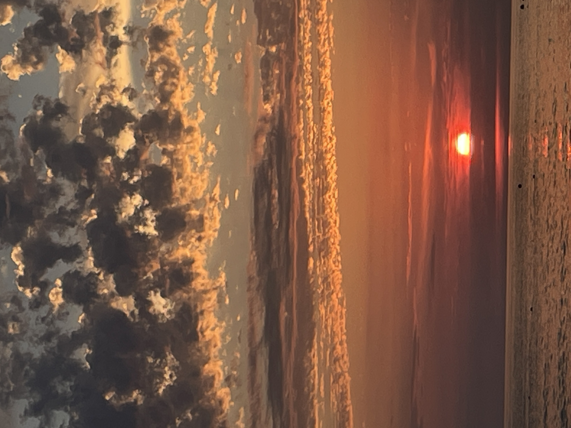 A vibrant sunset illuminates the sky with fiery oranges, reds, and purples, casting a warm glow over the calm, dark ocean. Wispy clouds, tinged with gold and dark gray, drift across the horizon, creating a dramatic and colorful display. The sun, a bright, glowing orb, is partially obscured by clouds, casting a shimmering reflection on the water's surface. The scene is tranquil and serene, evoking a sense of awe and wonder at the beauty of nature.