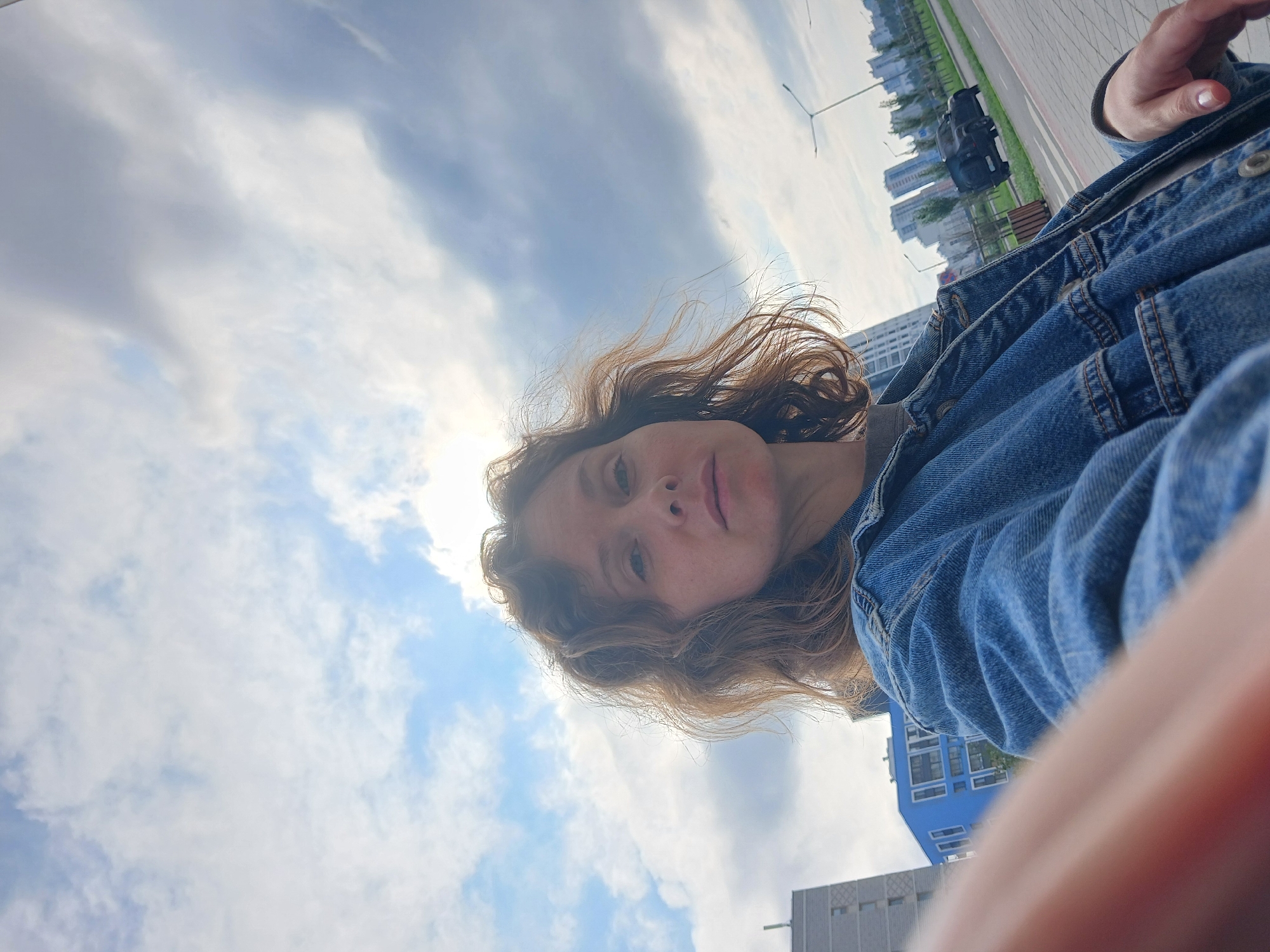 A woman with curly brown hair is taking a selfie outdoors. She is wearing a denim jacket. In the background, there are modern buildings, a street with a car, lampposts, and trees. The sky is cloudy with patches of blue. The image appears to have been taken during the daytime. Based on the architecture and overall aesthetic, the location is likely a modern urban development in a city, possibly on the outskirts.