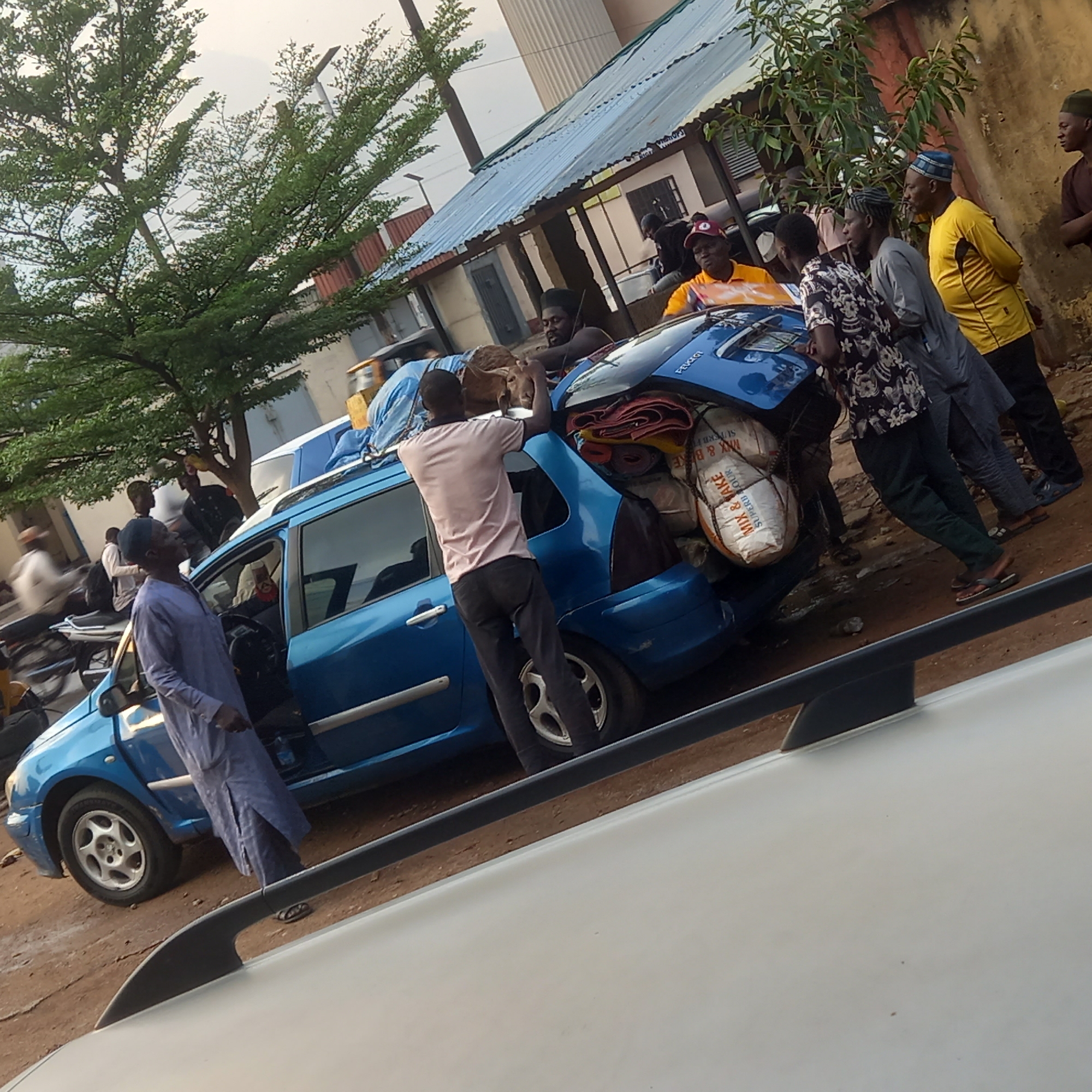 In Dadi Riba, Nigeria, a blue Peugeot 206 hatchback is heavily laden with goods, including bags of "Mix & Bake Super Flour," rugs, and at least one goat secured on top. A group of men are gathered around the vehicle, some assisting with loading. One man in a pink shirt is actively loading the vehicle, while another in a purple robe stands by the open driver's side door. Others are standing around. The car appears to be overloaded beyond its capacity, with the rear suspension visibly compressed. The scene is set outdoors, on a dirt or unpaved surface, adjacent to a building with a corrugated metal roof. The foreground is partly obscured by the roof of another vehicle.