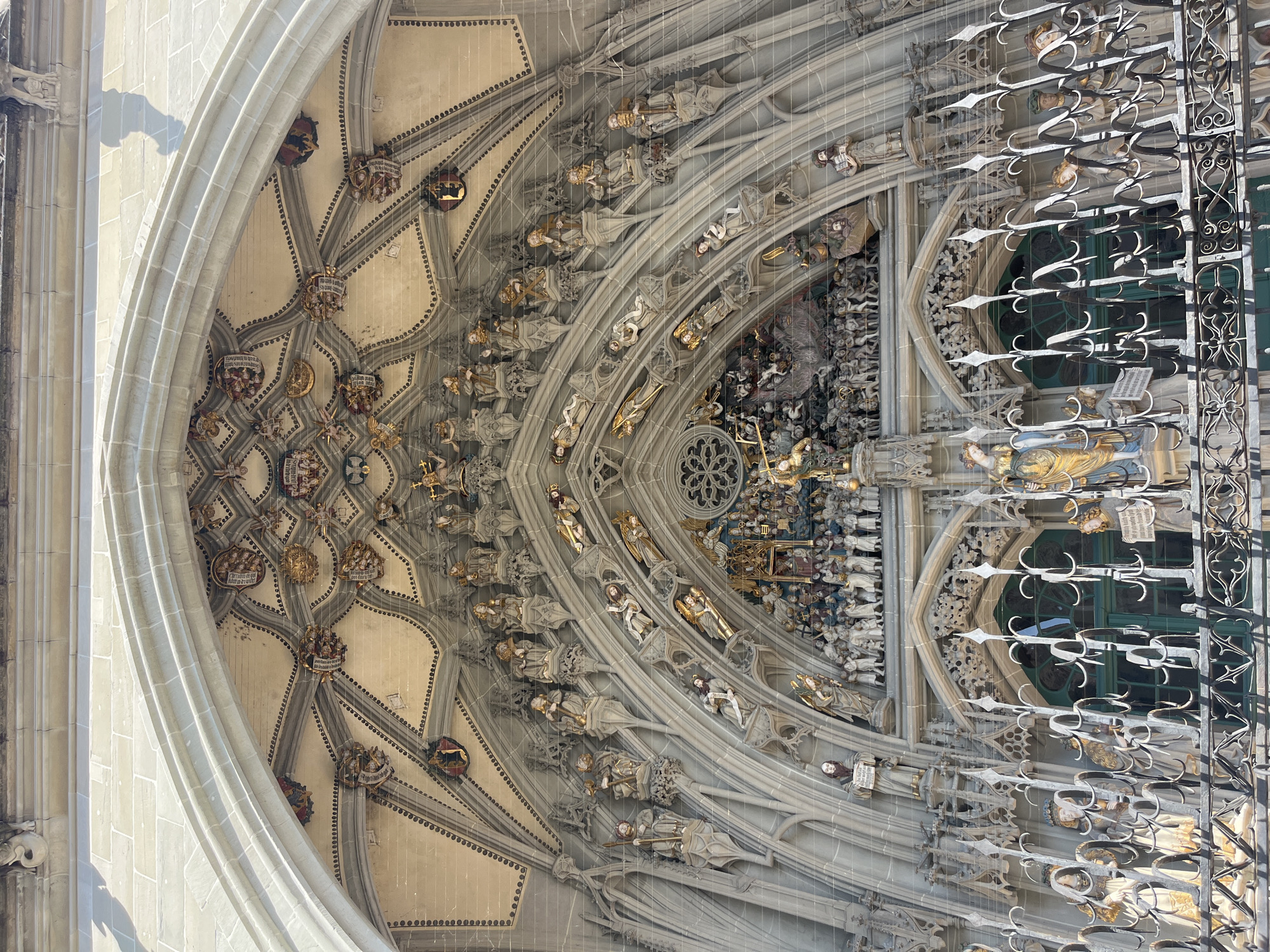 The image depicts the intricately carved facade of the Bern Minster in Bern, Switzerland. The photograph focuses on the grand Gothic entrance, showcasing a multitude of detailed sculptures adorning the portal. These sculptures include numerous figures of saints, angels, and other religious or allegorical characters, arranged in tiered niches and along the elaborate architectural framework of the arch. The carvings exhibit a rich level of detail, with figures depicted in various poses and holding symbolic objects. The stonework is weathered, indicating the age of the structure. The scene is illuminated by natural sunlight, casting shadows that highlight the three-dimensional quality of the sculptures. In the foreground, a wrought-iron fence with pointed finials runs across the bottom of the image, partially obscuring a view of more figures and a green-colored door or window. The overall impression is one of grandeur and historical significance, typical of major European cathedrals.