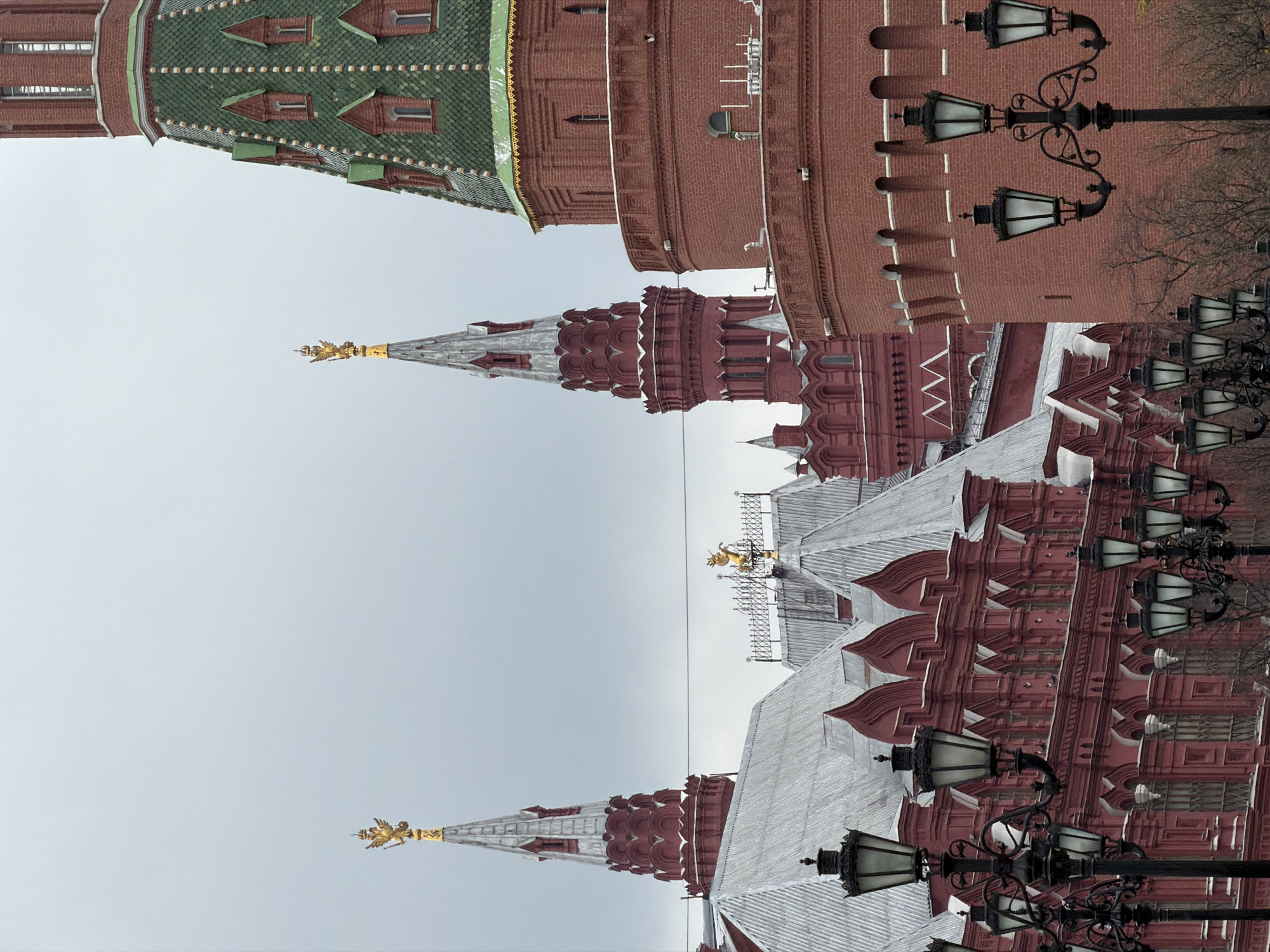 The image displays an expansive architectural complex, characteristic of the Moscow Kremlin, under an overcast sky. The scene is dominated by red brick structures and walls. On the left, a prominent tower features a distinct green tiled conical roof, punctuated by several small dormer windows. Towards the center, a narrower red brick tower with a grey conical roof culminates in a golden finial, depicting what appears to be a double-headed eagle, a national symbol of Russia.
To the right, extensive red brick walls with battlements (crenellations) are visible. Numerous ornate black lampposts, each with multiple lanterns, are positioned in the foreground and mid-ground, adding to the historical ambiance. The tops of various buildings within the complex showcase intricate red brickwork and white or grey roofs with decorative eaves. Another golden finial, similar to the one on the central tower, can be seen on a spire further to the right.
The weather is overcast, with a uniform light grey sky suggesting diffused light and no direct sunlight. The overall impression is one of a quiet, static moment. No people or activities are visible within the frame, and there is no discernible text present. The architectural style, including the red brickwork, specific roof designs, and the golden double-headed eagle finials, strongly identifies the location as Moscow, Russia.
