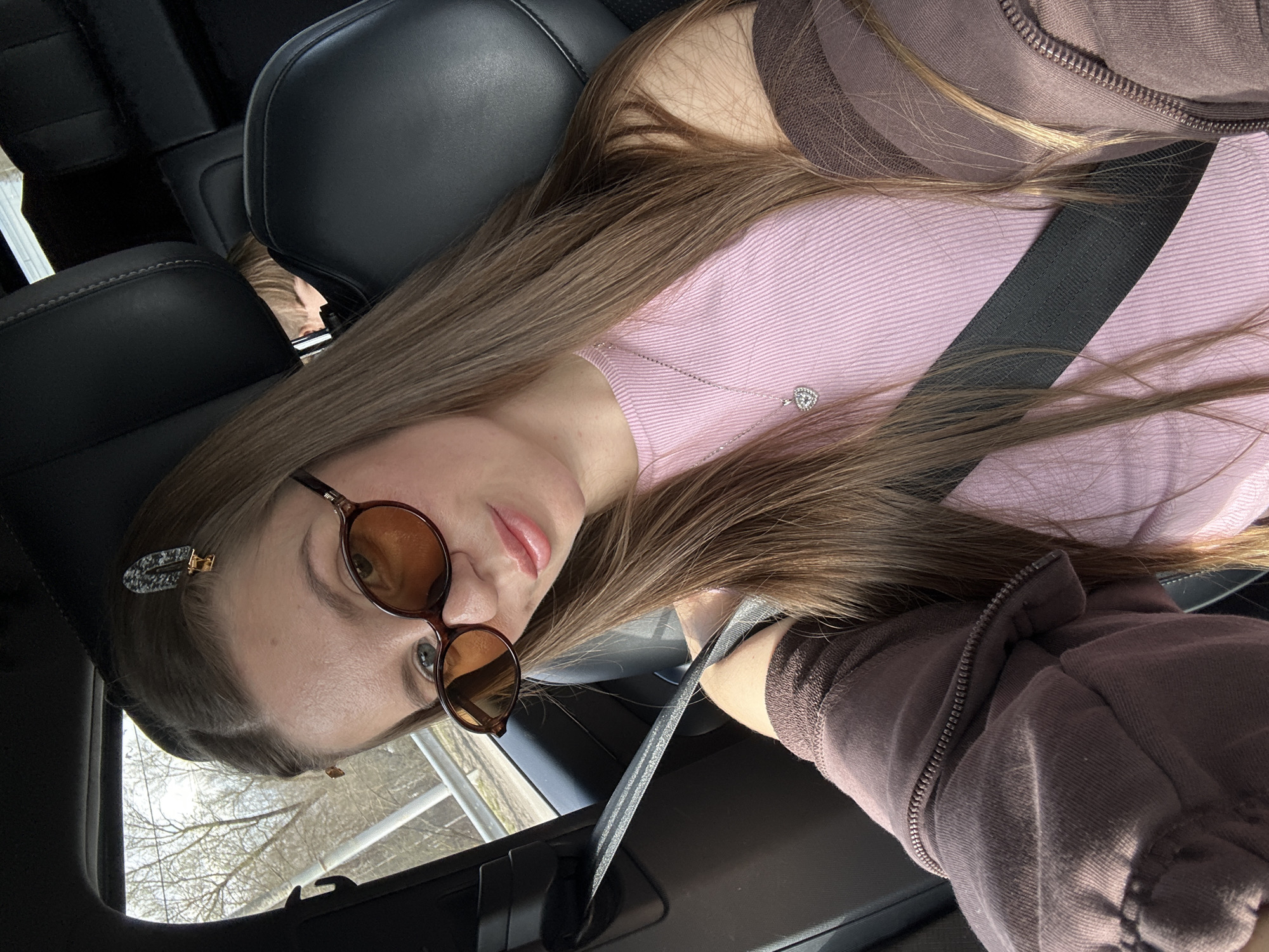 A young woman is captured in a close-up, rotated selfie taken inside a car, likely in Davydkovo, Russia. She has long, straight, light brown hair that flows over her shoulders and chest. She is wearing a light pink ribbed top under a dark brown or mauve zippered jacket. A silver necklace with a small, sparkly heart-shaped pendant is visible around her neck. Her face is partially obscured by round, brown-framed sunglasses with amber-tinted lenses, and a speckled hair clip is pinned to the side of her head. Her lips are slightly parted, conveying a neutral expression.

She is wearing a dark seatbelt across her chest. The car interior features dark leather seats and a black door panel. Bright daylight streams in through the window, indicating a daytime setting. Through the window, bare tree branches are visible against a bright sky, suggesting late winter or early spring. The scene depicts a casual moment of the woman inside the vehicle, possibly while in transit or parked. There is no visible text in the image.