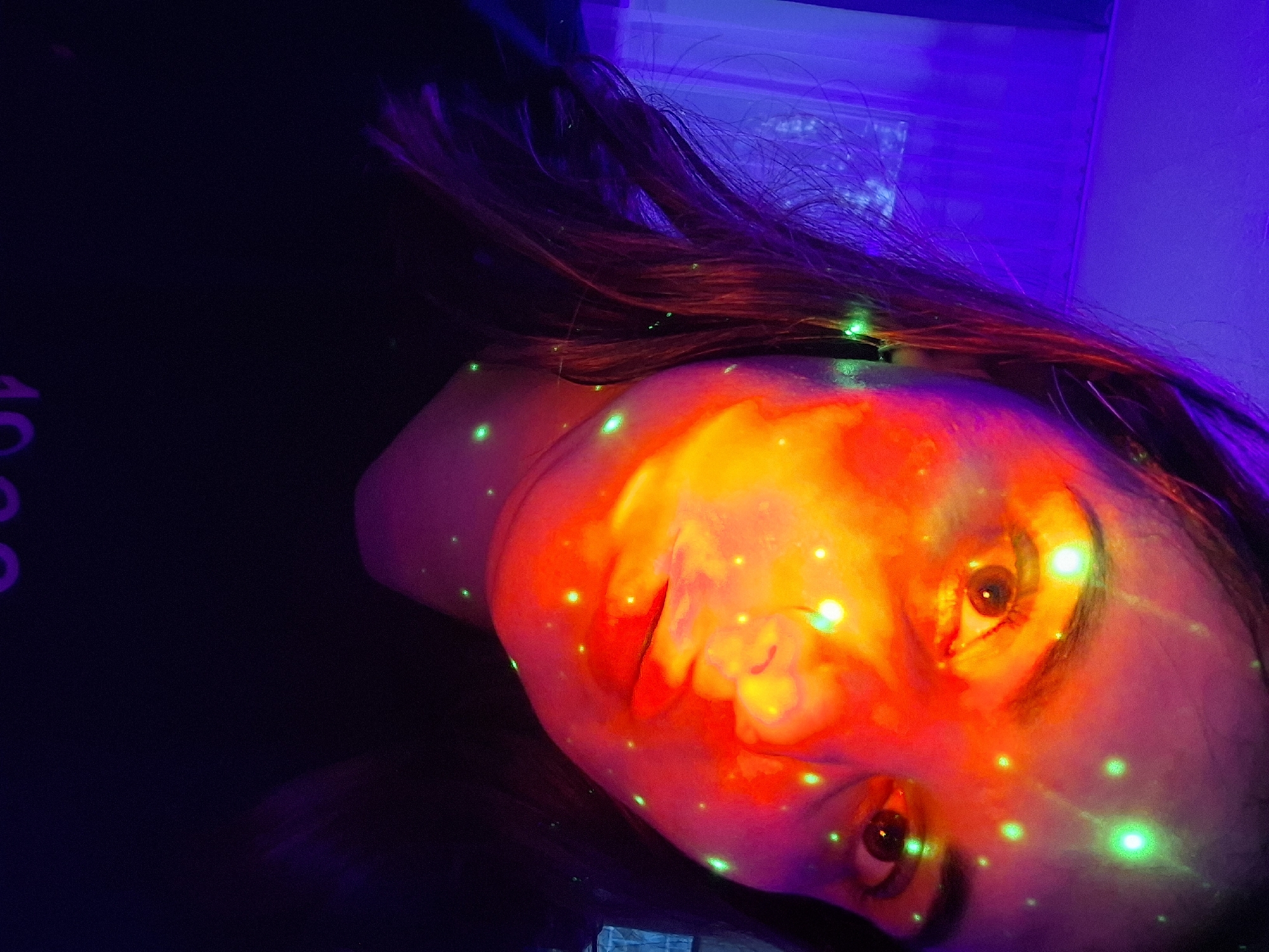 This image, captured in Chaikovsky, Russia, features a close-up portrait of a person, likely female, against a dark, neon-lit backdrop. The subject's face is angled sideways, dramatically illuminated by a vivid, intense red and orange glow emanating from an unseen light source. Numerous bright green, laser-like dots are scattered across her face, hair, and the dark background, creating a dynamic and ethereal effect.
Her dark hair cascades around her head, with hints of purple and blue ambient lighting visible in the upper right portion of the frame, suggesting a larger light installation or room lighting. The overall setting appears to be an indoor space, such as a nightclub, party, or artistic light installation, indicated by the artificial, high-contrast lighting and dark surroundings, consistent with a nighttime environment. The person's eyes are open and directed towards the viewer, conveying a calm or possibly captivated expression amidst the light display. On the far left of the dark background, partial purple numbers, resembling "10" or "100," are faintly visible. The striking interplay of vibrant colors and intricate light patterns creates a surreal and engaging visual experience.