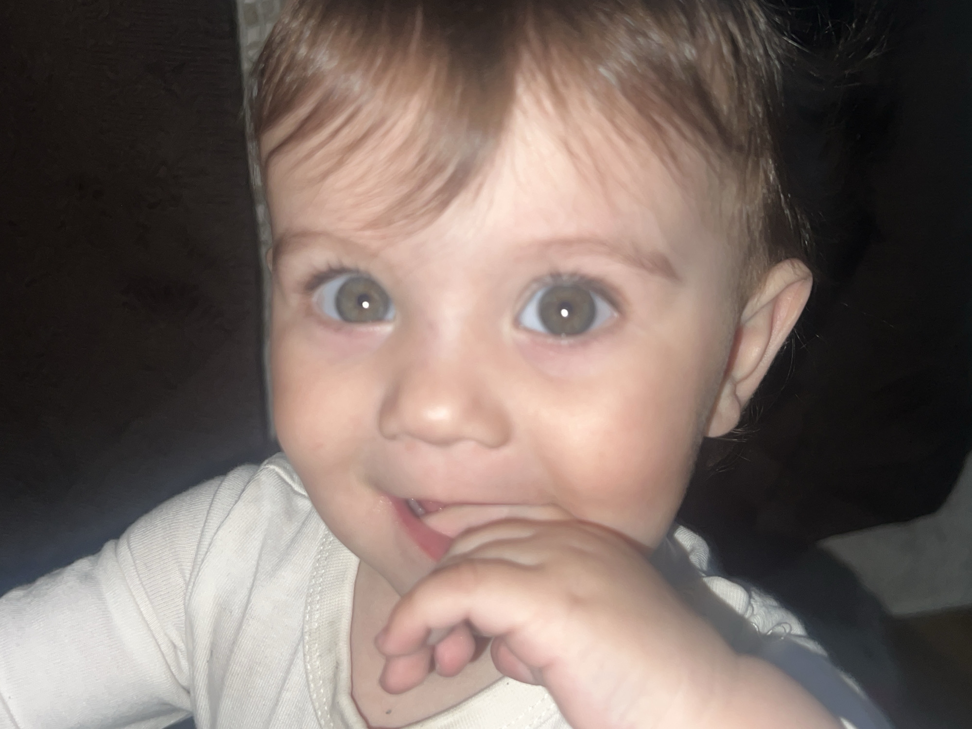 A close-up shot focuses on a fair-skinned baby with light brown hair, gazing directly at the camera with wide, inquisitive brown eyes. The baby's fingers are touching their mouth. The baby is wearing a white, long-sleeved shirt. The background is mostly dark and out of focus, obscuring details. There is no indication of Elena, Bulgaria.