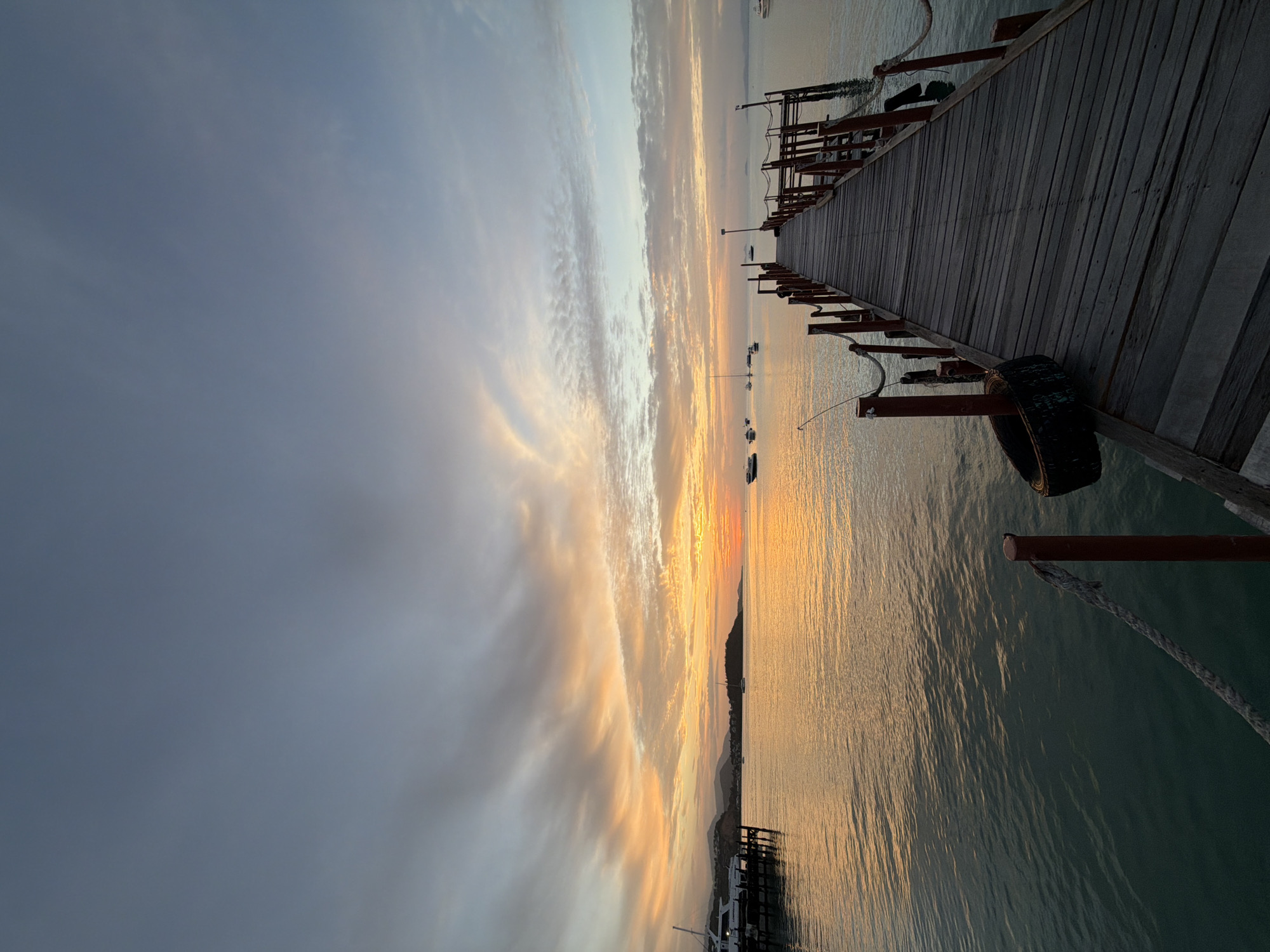 This is a photograph of a wooden pier extending into the ocean at sunset in Ko Samui District, Thailand. The sky is filled with colorful clouds in shades of orange, yellow, and blue, reflecting on the calm water. Several boats and sailboats are docked or anchored in the distance. The pier itself is made of weathered wooden planks and has red posts and ropes along its sides. A worn tire is attached to one of the posts near the beginning of the pier. The scene evokes a peaceful and serene atmosphere, typical of a tropical island sunset.