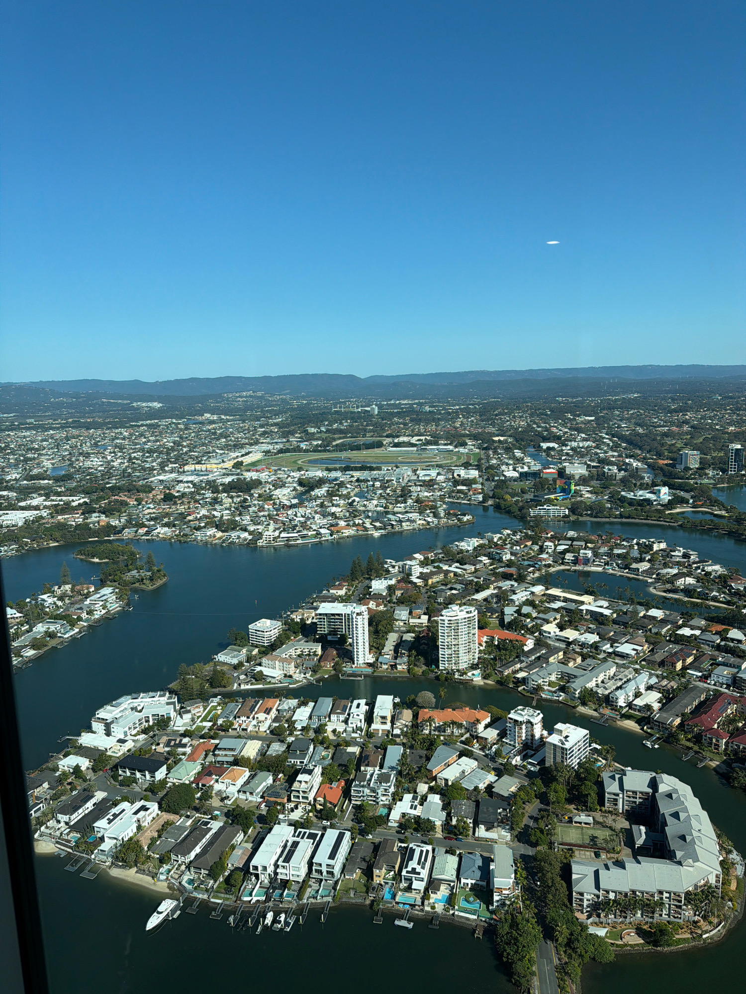 This is an aerial photograph of the Gold Coast, Australia, likely taken from a helicopter. The image showcases a densely populated urban and suburban landscape characterized by a network of waterways and canals. Numerous residential buildings, ranging from multi-story apartment complexes to single-family homes with private docks and swimming pools, line the water's edge. Vegetation, including trees and manicured lawns, is interspersed throughout the residential areas. In the background, a broader urban sprawl extends towards a distant, hazy horizon with rolling hills. A faint white elliptical object is visible in the upper right quadrant of the sky. The scene is under a clear, bright blue sky, suggesting a sunny day.