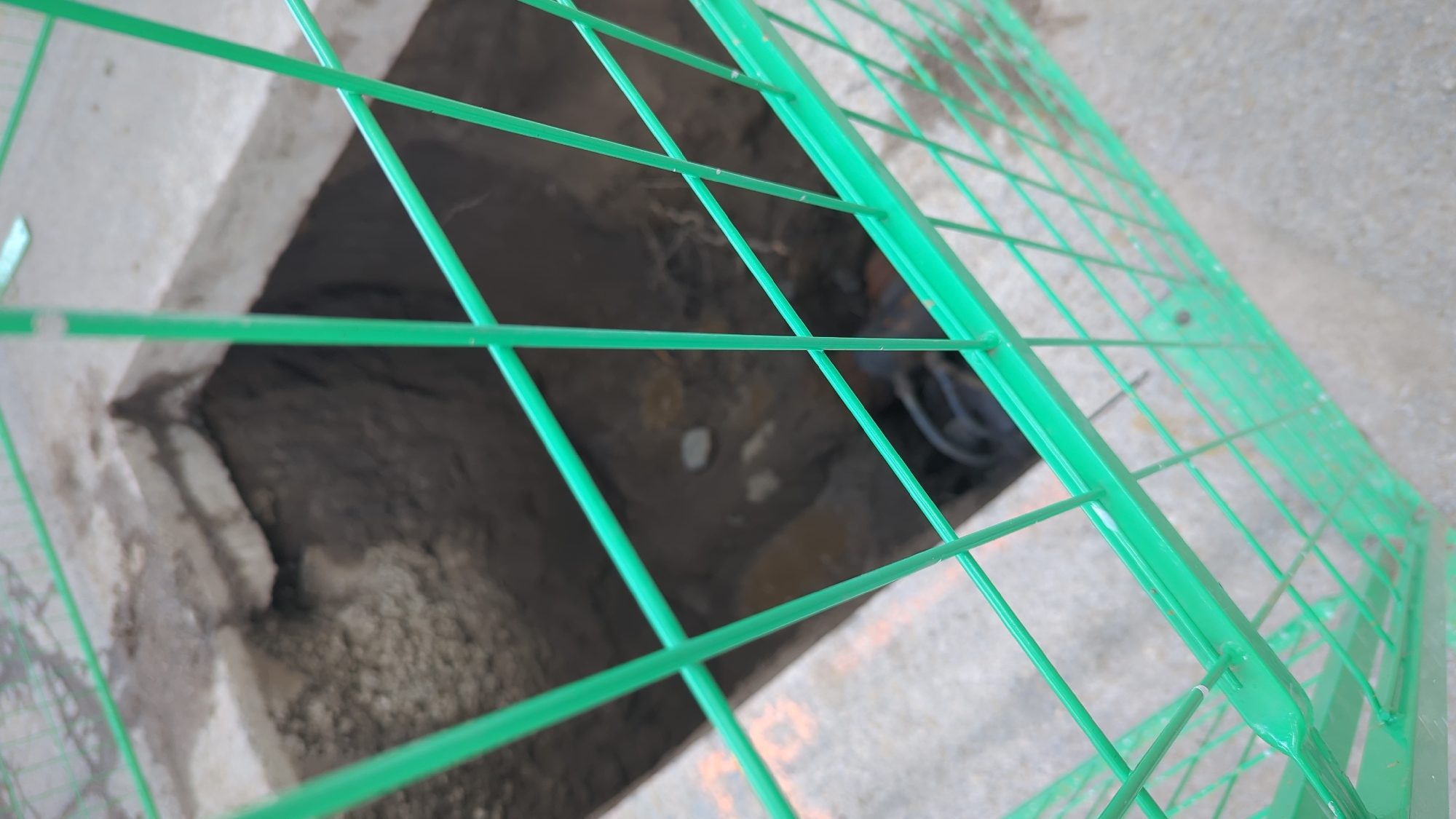 The image shows a construction site in Montreal, Canada. A hole has been dug in the ground, and it is surrounded by a green metal fence. The hole appears to be deep and dark, with some debris at the bottom. The fence is made of vertical and horizontal bars, and it is blocking access to the hole. The ground around the hole is concrete, and there are some orange markings on the concrete. The scene appears to be outdoors, and the lighting suggests it is daytime. There are no people visible in the image.