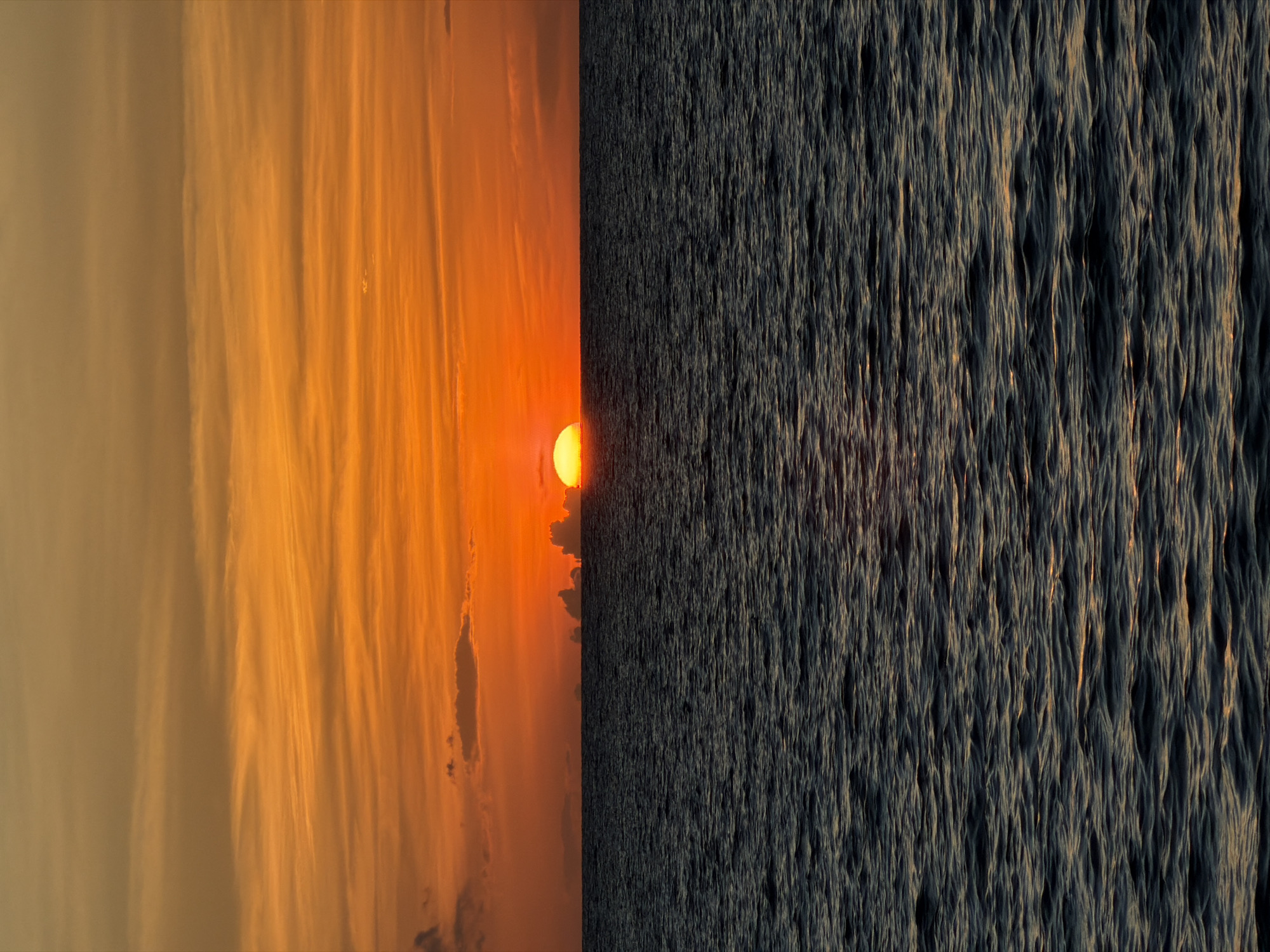 This image is a diptych, with the left side showing a vibrant sunset over the ocean and the right side displaying the textured surface of the ocean. On the left, a bright orange sun is setting, casting a warm glow on wispy, streaky clouds painted in shades of orange, yellow, and pink. The sky is dramatic and colorful, indicative of a beautiful sunset. The ocean below is calm, reflecting the sky's hues.
The right side of the image presents a close-up view of the ocean's surface. The water appears choppy or rippled, creating a pattern of dark, elongated waves or troughs that run vertically. Sunlight catches the crests of these waves, highlighting them with a golden or amber light, contrasting with the deep blue-gray of the water. This side of the image feels more textured and dynamic, showcasing the movement and form of the water itself.
The overall impression is one of nature's beauty, capturing a moment of transition between day and night. The setting is clearly a coastal environment, given the presence of the ocean and the sunset. The time of day is sunset, as evidenced by the sun's position and the colors in the sky. The weather appears clear, allowing for unobstructed sunset views. No people, objects, or text are visible in the image.