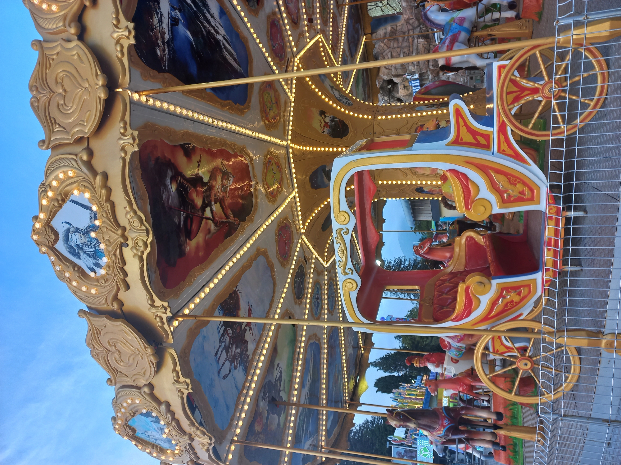 The image shows a carousel ride in Sochi, Russia. The carousel features a golden canopy adorned with paintings and illuminated by strings of lights. A carriage-shaped seat, painted in red, yellow, and blue, is visible on the right side of the carousel. The carousel is surrounded by a fence. The background includes trees and a blue sky.