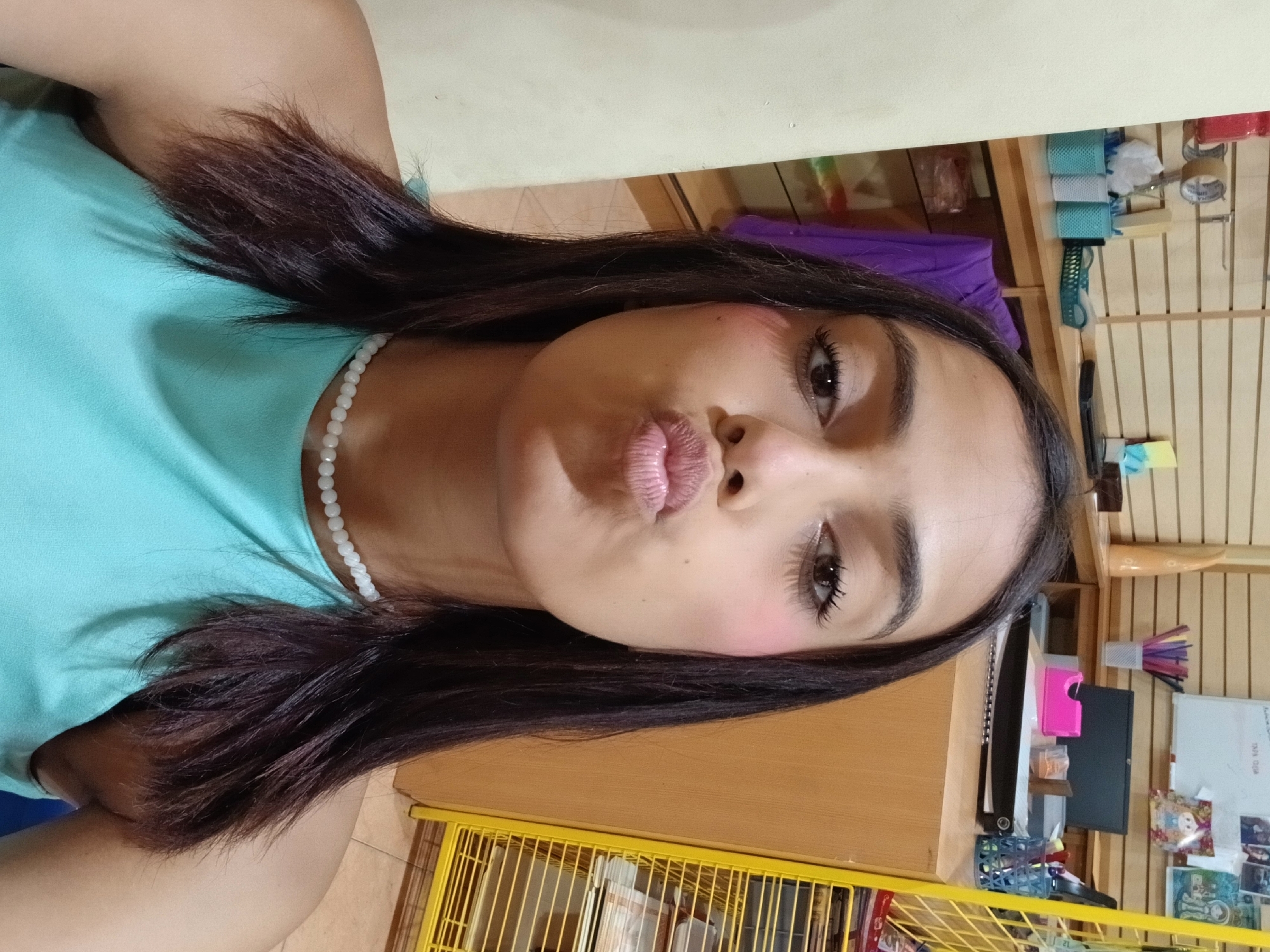 The image shows a medium shot of a young woman with dark hair, puckering her lips and looking at the camera. She is wearing a light blue sleeveless top and a white beaded necklace. Her cheeks have a light pink blush. The background appears to be an indoor setting, possibly a shop or office, with shelves displaying various items, including stationery and what looks like merchandise. There is a desk with a laptop, a pink tape dispenser, and pens in a holder. To the right, a yellow metal shelving unit is visible. The lighting is artificial and suggests an indoor environment. While the image captures a personal moment, the broader context suggests a commercial or retail setting, potentially in a town like Juan Griego, Venezuela, known for its markets and shops.