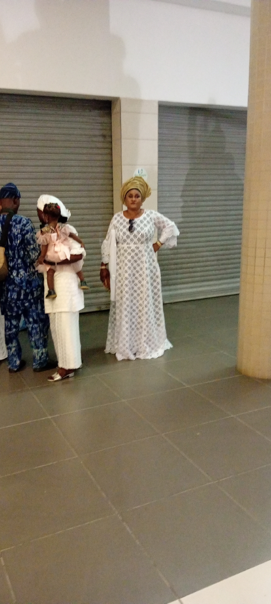 In Gandu Albasa, Nigeria, a woman in a white, intricately patterned dress and a gold headwrap stands with her hand on her hip, facing forward. She wears sunglasses on her head and has a gold bracelet on her left wrist. To her left, a woman in a white dress and headwrap holds a young child dressed in a pink ruffled dress. The child faces away from the camera, with their back to the viewer. To the left of the woman holding the child, a man in a dark blue and white patterned traditional outfit faces away from the camera, with his left arm across his body. The group is positioned in an indoor setting with a tiled floor and large, closed metal shutters in the background. A beige pillar is visible on the right side of the image.