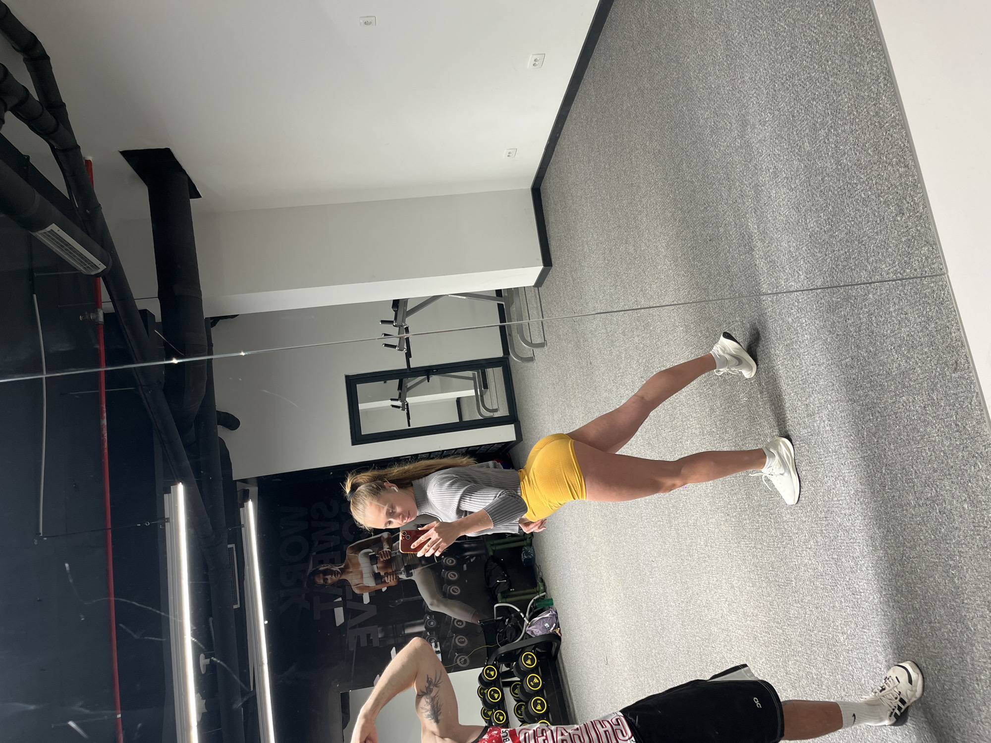 A woman in yellow shorts and a grey long-sleeved shirt stands in front of a large mirror in a gym. She is holding a phone, presumably taking a selfie or video. The reflection in the mirror shows a weight rack with dumbbells, fitness equipment, and a black wall with graphics. Another person is partially visible at the bottom of the mirror, wearing black shorts and a red shirt. The gym has a gray floor and white walls. Black piping is visible on the ceiling. The location is likely Boreti, Montenegro.