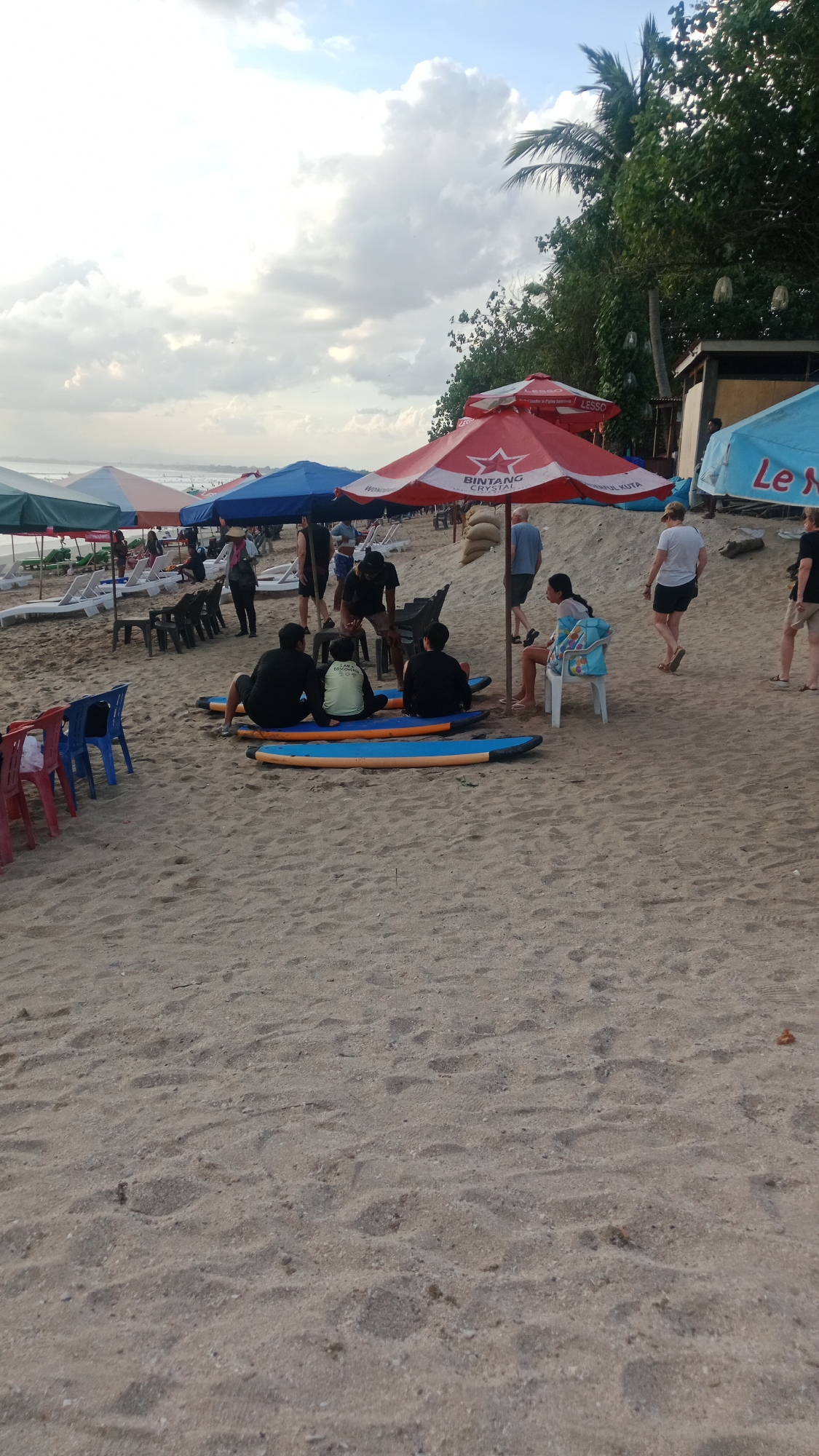 The image captures a beach scene in Kuta, Indonesia, characterized by fine sand under a partially cloudy sky. Several beach umbrellas are set up, including a prominent red one with the "Bintang Crystal" logo, indicating a likely location near a beachfront establishment.
In the foreground, three individuals are seated on surfboards on the sand, appearing to be taking a break or waiting. Two of them are wearing dark shirts, and one is wearing a lighter colored shirt. Nearby, a woman sits on a white plastic chair with a blue backpack, also under an umbrella. Another person, possibly a server or instructor, stands near them.
Further back, a group of people are visible near rows of lounge chairs and umbrellas. Some are standing, while others are seated. Several individuals are walking along the beach. To the right, a person in a black t-shirt and shorts is walking away from the camera, and to their left, another person in a white t-shirt and shorts is walking towards the viewer. The background shows trees and a building, suggesting the presence of beachfront infrastructure. The overall atmosphere is relaxed, with a mix of leisure and activity characteristic of a popular beach destination.