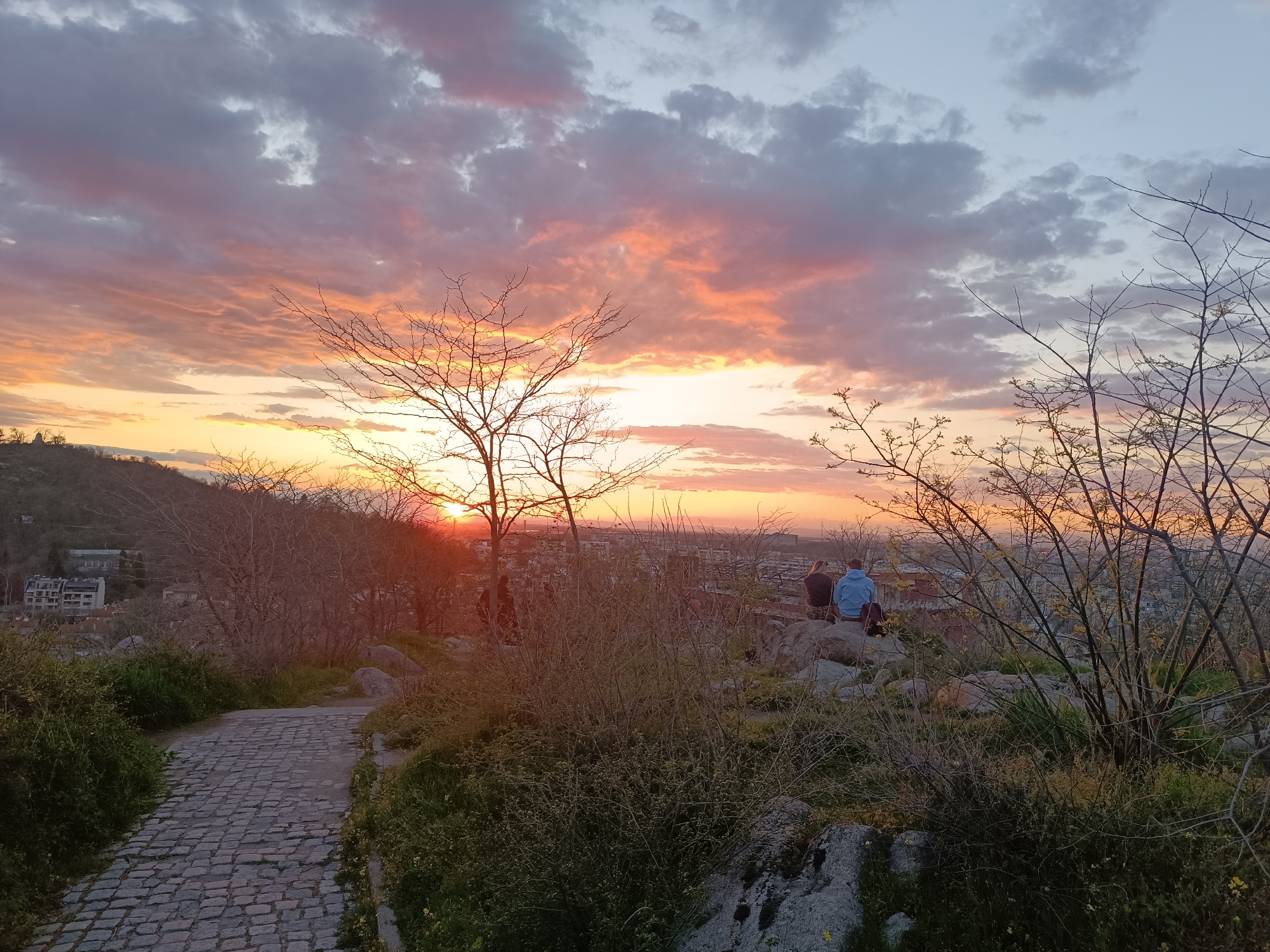 A wide-angle shot captures a sunset in Unknown, Unknown, with a cobblestone path leading towards the foreground and a landscape bathed in warm hues. Two people, one in a blue sweatshirt, sit on rocks in the middle distance, gazing towards the city visible through bare branches. The sky is filled with orange, pink, and gray clouds. Bushes and trees occupy the foreground.