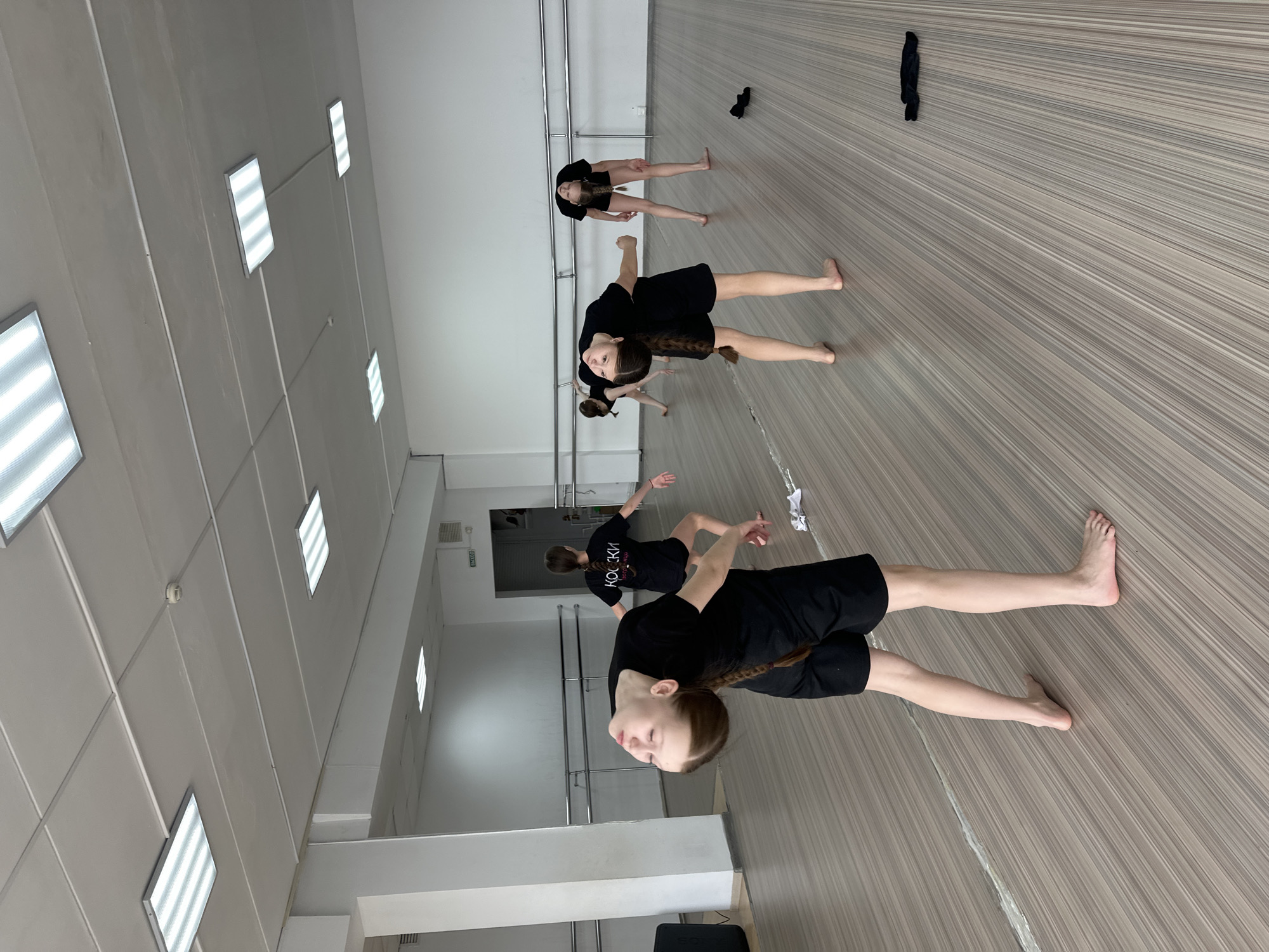 The image shows a dance studio in Votkinsk, Russia. Several young girls, dressed in black attire and with long braids, are practicing a dance routine. They are barefoot on a wooden floor in a room with white walls and ceiling, illuminated by fluorescent lights. A ballet barre runs along the wall. There is a black garment lying on the floor. A mirror is visible on one side of the room.