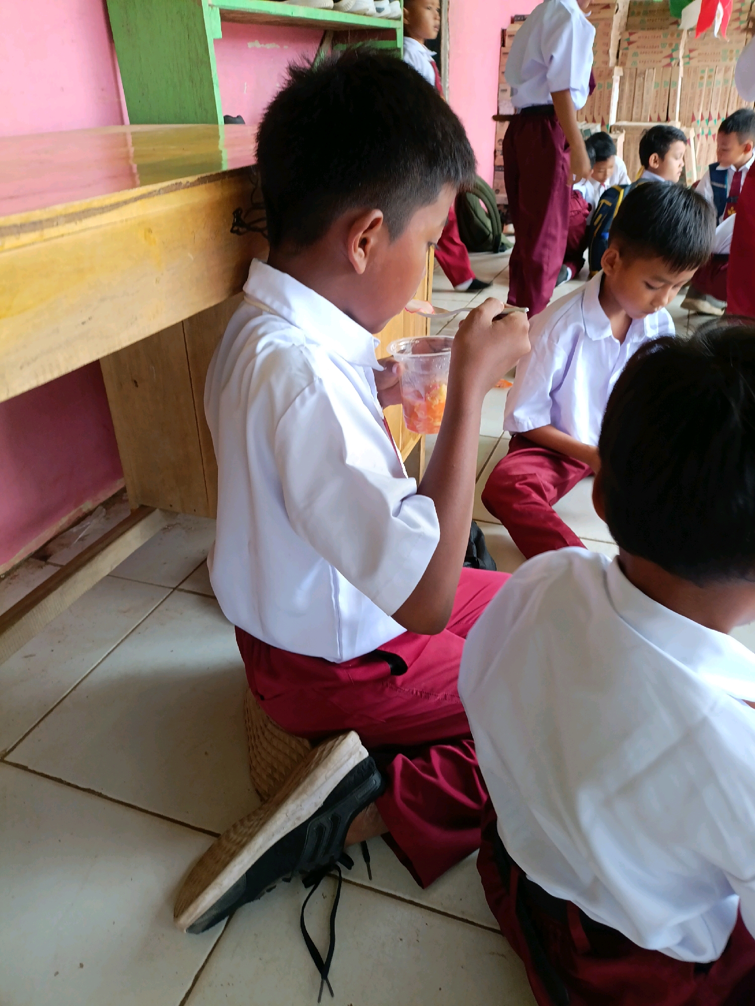 This image depicts a group of young students, likely in a school setting in Sumberejo, Indonesia, wearing white shirts and maroon pants. The foreground features a boy on the right, seen from behind, with a white shirt and maroon pants. To his left is another boy, also in a white shirt and maroon pants, sitting and eating from a clear plastic cup with a spoon. His left arm is visible, holding the cup. To the left and behind these two boys, a wooden structure with a yellow top and pink and green sides is visible, possibly a shelf or bench. In the background, several other students are present, also in similar school uniforms. Some are standing, and one is seated. The environment appears to be indoors, with tiled flooring and a wall with various items on shelves in the background, including boxes and what appear to be decorative flags or banners. One student in the background is wearing a blue vest over their white shirt. The lighting suggests an indoor space, possibly with natural light entering from an unseen source.