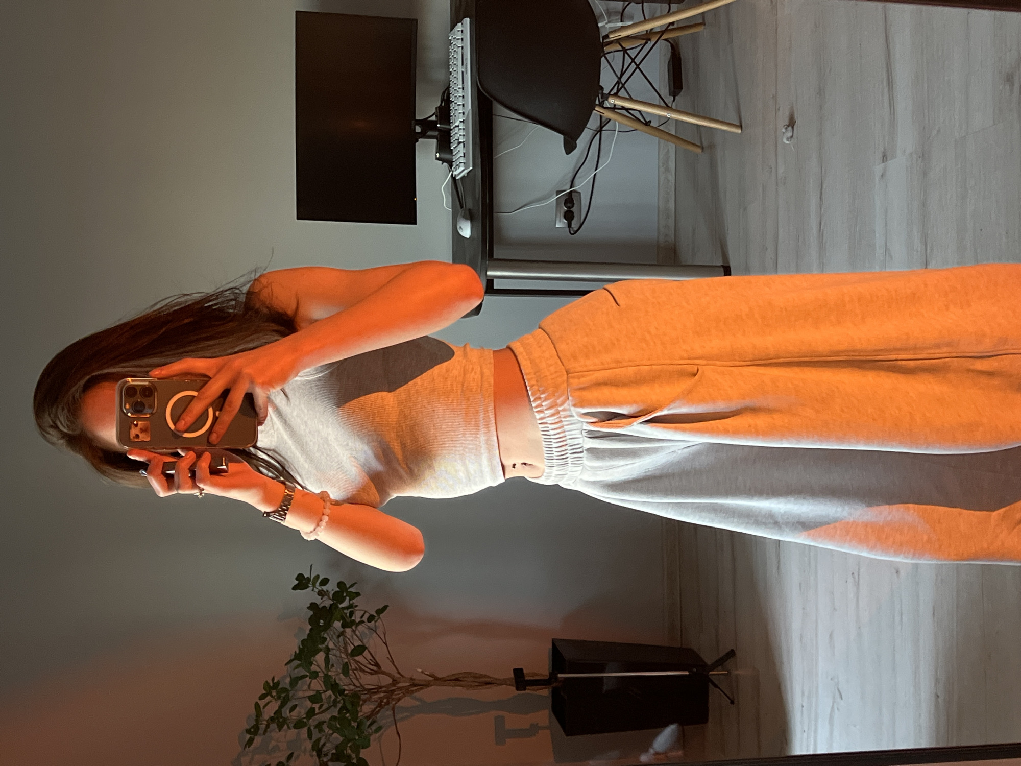 In an indoor setting, a young woman is taking a mirror selfie. She is wearing a light gray cropped tank top and light gray sweatpants. Her hair is dark brown and flows down her shoulders. She has a navel piercing. On her left wrist, she wears a watch and a beaded bracelet. In her hands, she holds a smartphone, angled to capture her reflection.
The background suggests a home office or a room with a desk. A black computer monitor sits on a black desk, with a white keyboard and mouse. A black chair with wooden legs is positioned at the desk. To the left of the woman, a potted plant with sparse foliage stands in a tall black planter. The room is illuminated by warm, orange-toned lighting, casting long shadows. The floor appears to be light-colored laminate. The image is taken in Ternopil, Ukraine.