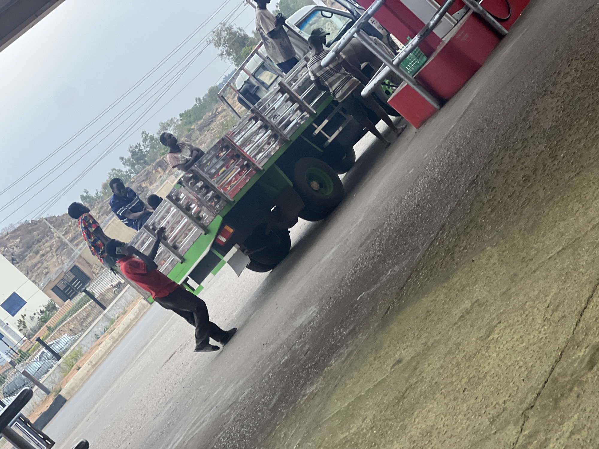The image presents an outdoor scene centered around a green flatbed truck with a white cab, surrounded by several men, in what appears to be Gura Top, Nigeria. The photograph is notably tilted, creating a diagonal horizon.
In the foreground, a man in a red shirt and black pants is leaning forward, holding onto a metal railing or a similar structure, with another man in a patterned shirt partially visible behind him. The green flatbed truck is the main object, featuring a protective metal railing around its bed. Several men are engaged around the truck; one stands in the truck bed near the cab, wearing a light-colored shirt, while another in a striped shirt and a dark cap stands on the ground next to the truck's front passenger side. More individuals are visible on the truck bed, suggesting a loading, unloading, or transit activity.
To the right, red structures resembling fuel pumps or a station kiosk are visible, including red cabinet-like elements and metal railings. The ground beneath the truck and figures is paved with asphalt or concrete, appearing damp in certain areas. In the mid-ground and background, the landscape includes rocky hills or mountainous terrain with scattered trees and vegetation under a bright, hazy sky. On the far left, a building with a white facade and a dark fence is partially visible, with a parked car behind the fence. Overhead, power lines span across the sky.
The scene depicts a daytime setting, characterized by soft, diffused lighting consistent with an overcast sky or a specific time of day like morning or late afternoon. There are no strong shadows present. No distinct text is clearly legible on any of the objects or structures within the frame. The overall atmosphere suggests a casual or work-related gathering around the truck at a roadside stop.