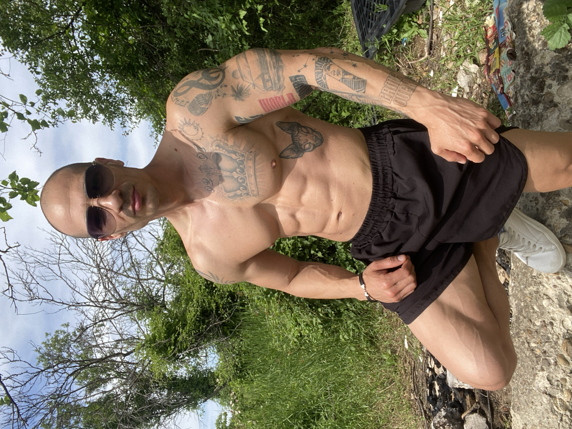A shirtless man with a shaved head, sunglasses, and multiple tattoos poses outdoors. He wears black shorts and white sneakers. The background includes green foliage and a glimpse of a sky with tree branches. The location appears to be a natural area, likely in or near Pleven, Bulgaria, given the city specification. There is a stone or concrete surface at his feet. He is touching his shorts.