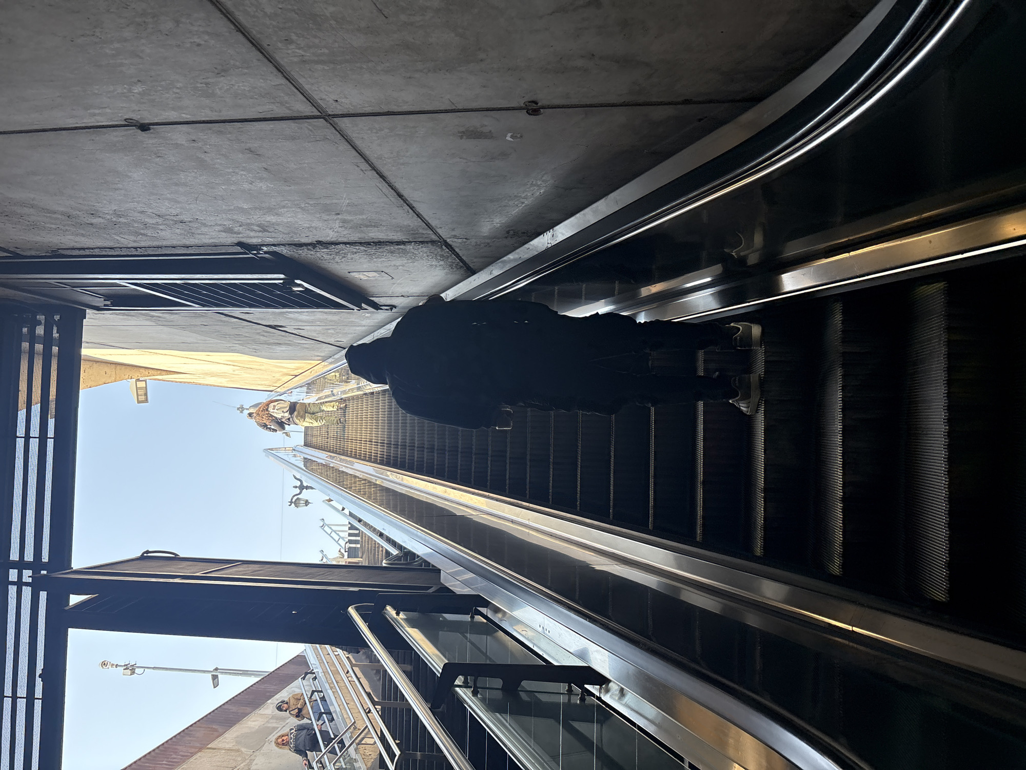 The image shows an upward-facing view of an escalator in a modern, possibly underground, transit station. The escalator is predominantly black with silver-toned handrails. A person, wearing a dark hooded jacket and dark pants, is ascending the escalator, positioned centrally in the frame and facing away from the viewer. Their back is to the camera. To the left, another set of escalators or stairs with metal railings is visible, with a few people in the distance ascending. The surrounding architecture features concrete walls with visible seams and some yellow-colored structures higher up, suggesting a contemporary urban design. The sky is bright blue and visible through an overhead opening, indicating it is daytime. The overall environment is clean and functional, typical of public transportation infrastructure. The location is identified as Santiago, Chile.