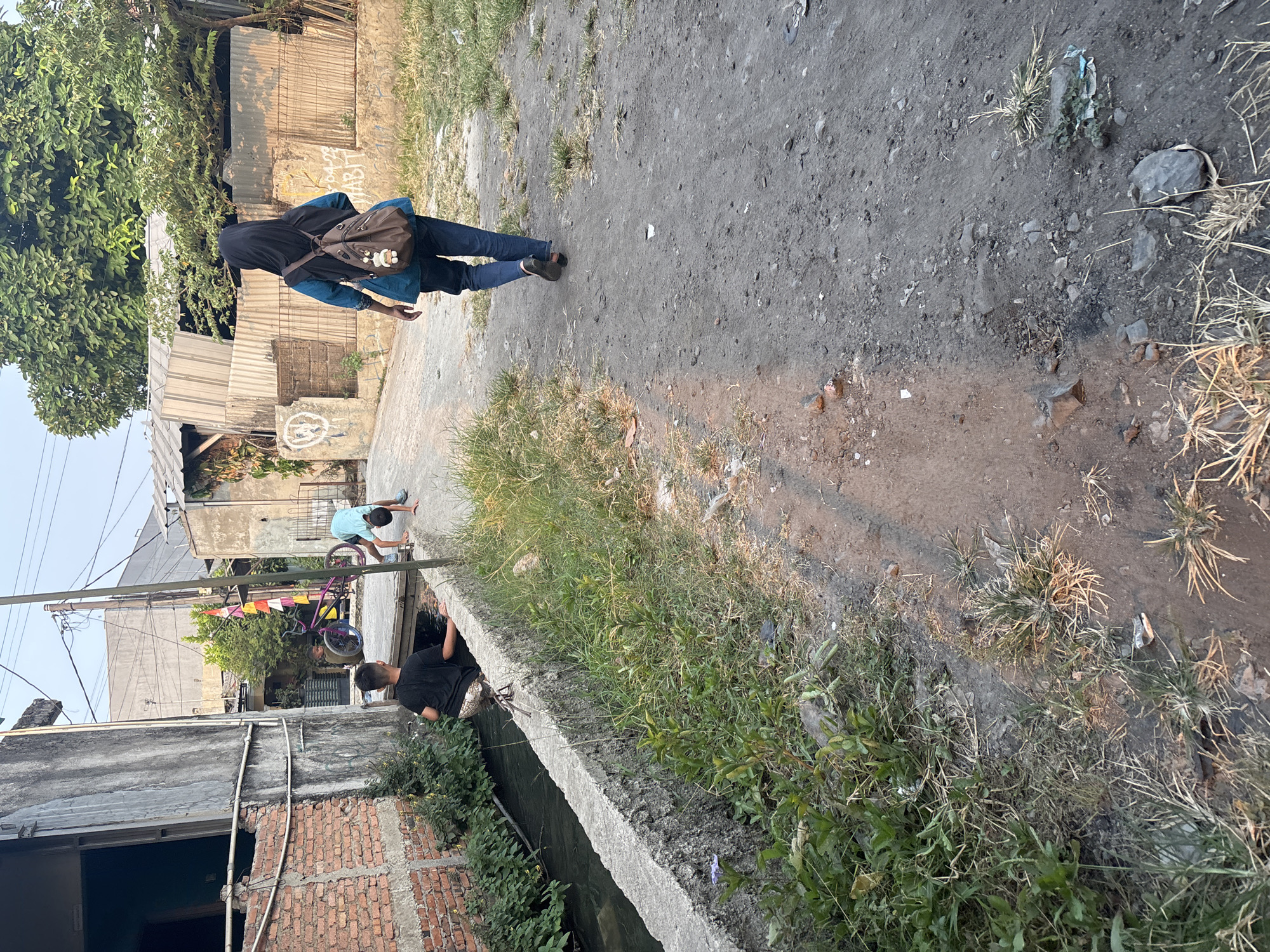 The image depicts a scene in Telagamurni, Indonesia, featuring a narrow, somewhat overgrown dirt path adjacent to a concrete-lined canal. Two young boys are present near the canal. One boy, wearing a black t-shirt and patterned shorts, is partially in the water, appearing to interact with the canal. Another boy, wearing a light blue t-shirt and shorts, is in a dynamic pose on the path, with his hands on the ground as if doing a cartwheel or stretching. A purple bicycle with blue rims is parked nearby. In the background, several simple buildings with corrugated metal and brick construction are visible. A young person wearing a black hijab, blue top, and jeans, carrying a brown backpack, walks away from the viewer down the path. The environment is characterized by dry grass, scattered debris, and a general sense of an urban fringe area.