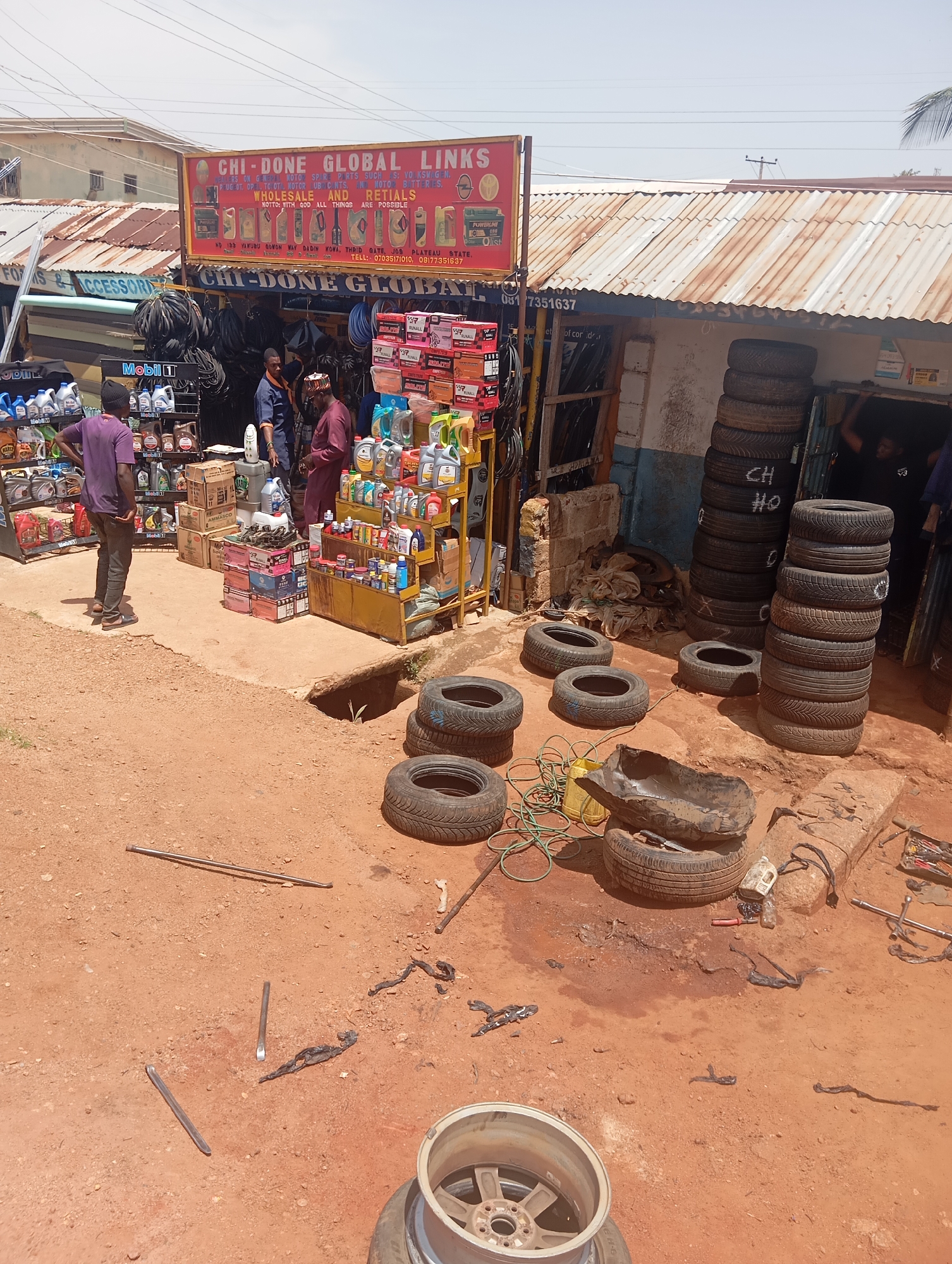 The image depicts an outdoor scene at a tire and auto parts shop in Jos, Nigeria. The shop has a red and yellow sign that reads "CHI-DONE GLOBAL LINKS WHOLESALE AND RETIALS" and includes a phone number. Stacks of tires are visible, both on display and in piles on the ground. Shelves are stocked with various automotive fluids, including engine oil and coolants, with prominent branding such as "Mobil 1". Several people are present, including a man in a purple shirt and dark pants standing near a display of oil, and two men in traditional Nigerian attire engaged in what appears to be a transaction or discussion. The ground is a mix of dirt and concrete, with various tools and discarded tire parts scattered around, suggesting active repair work. The environment appears to be a busy marketplace or street-side establishment.