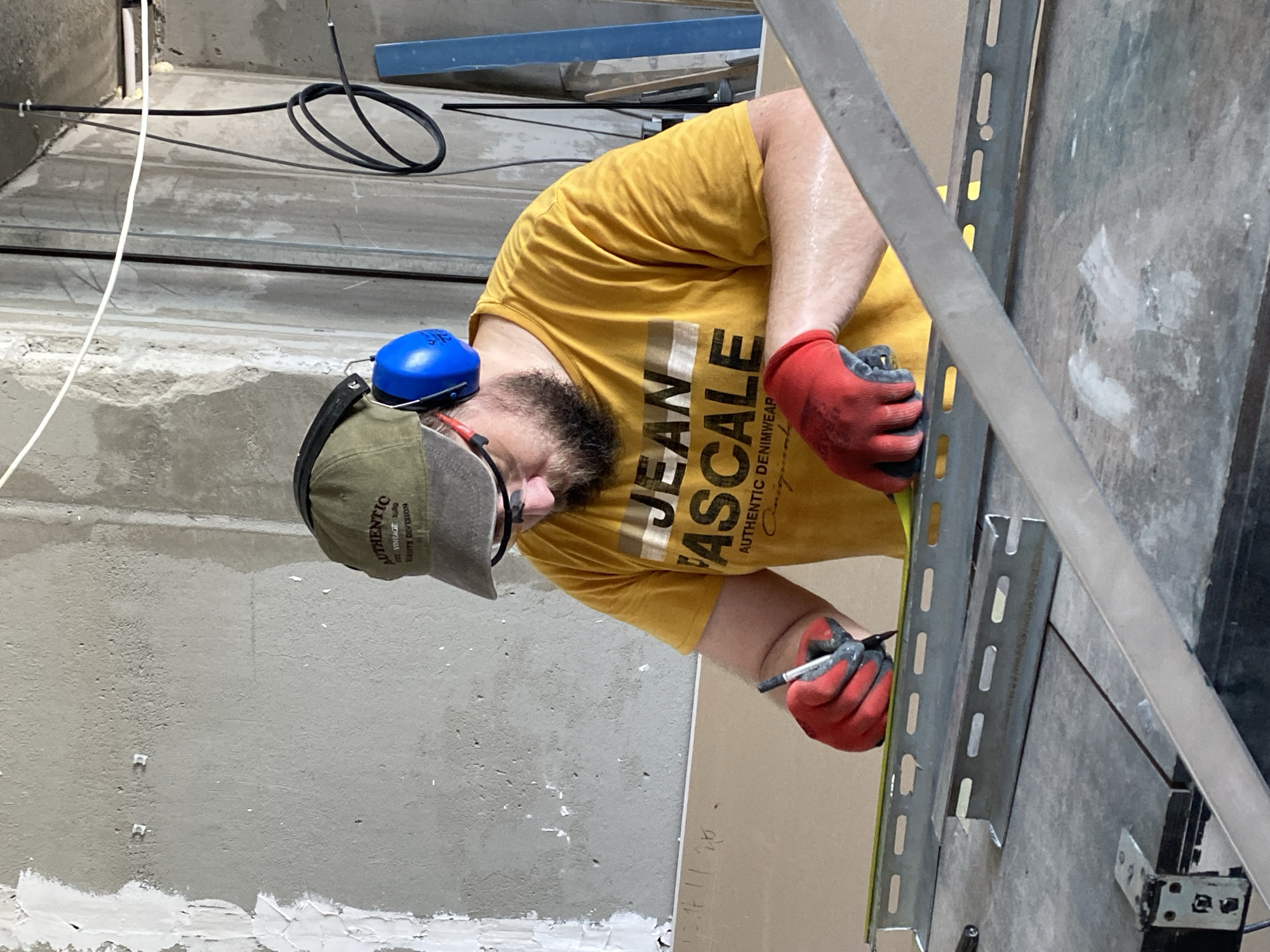 The image depicts a man wearing a green cap with "AUTHENTIC" text, blue earmuffs, and red work gloves. He is also wearing glasses and a yellow t-shirt with "JEAN ASCALE AUTHENTIC DENIMWEAR" printed on it. The man is in a construction or workshop environment, with a grey concrete wall in the background. He is actively engaged in measuring and marking a piece of metal railing on a workbench. The workbench is cluttered with various tools and materials, suggesting ongoing construction or repair work. The scene appears to be indoors, possibly a workshop or a room under renovation. The presence of construction materials and tools indicates a focus on construction or DIY activities. Without further information or visual cues linking this specific scene to Batumi, Georgia, it's impossible to confirm its location within the city.
