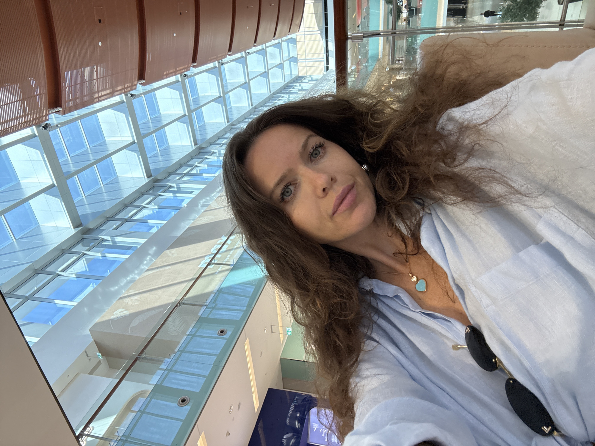 The image shows a woman with long, wavy brown hair and blue eyes taking a selfie indoors. She is wearing a light blue button-down shirt, a necklace with a turquoise heart pendant, and sunglasses tucked into her shirt. The background suggests an indoor public space with a modern architectural design, featuring a glass ceiling and multi-level walkways, indicative of a shopping mall. The overall lighting is bright, with natural light filtering through the glass ceiling. The architectural style and design elements, such as the glass facades and suspended walkways, are characteristic of upscale shopping destinations found in Dubai, United Arab Emirates, such as the Dubai Mall or Mall of the Emirates.