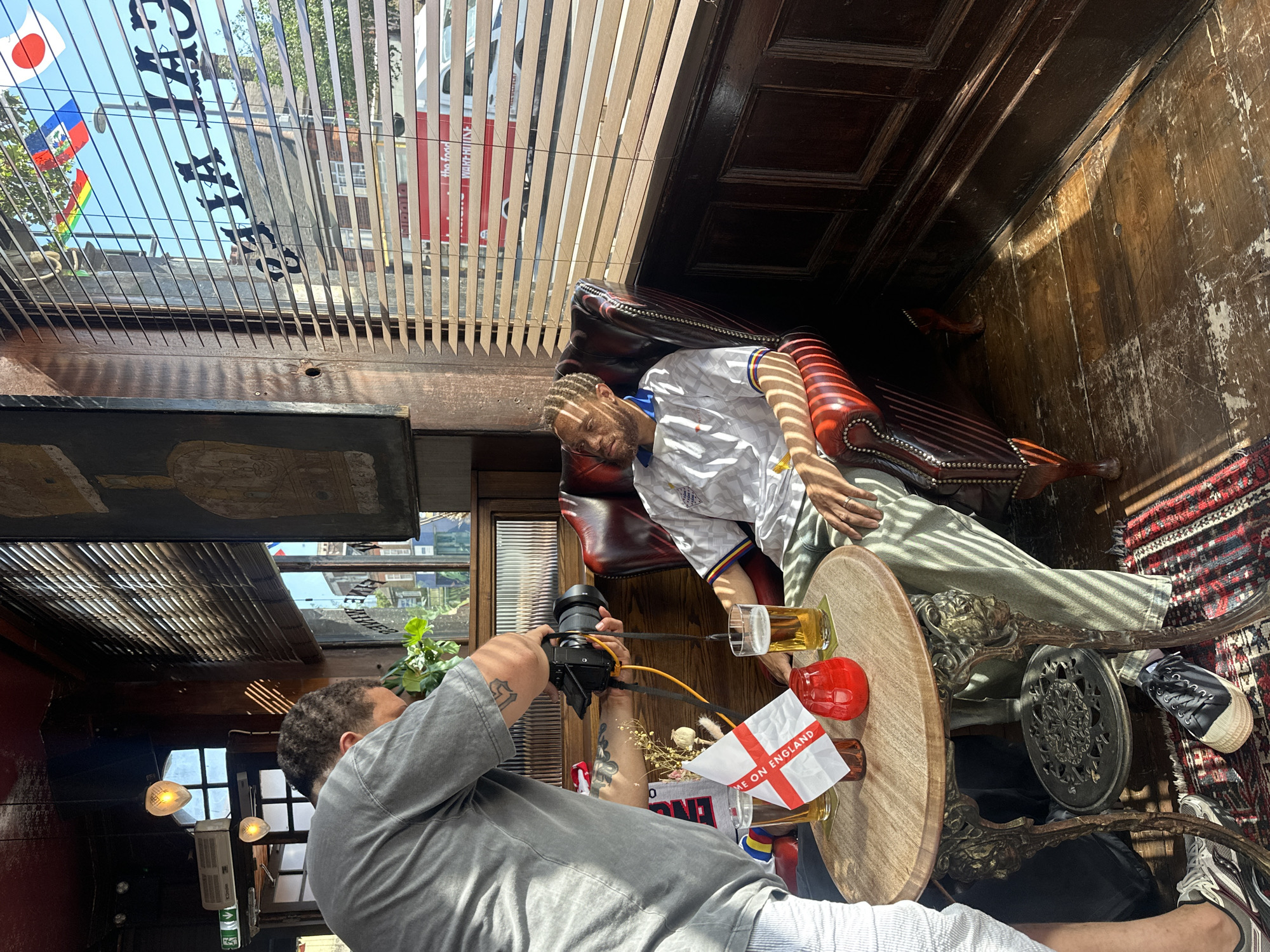A man is taking a photo of a mannequin sitting in a leather armchair inside a pub. The mannequin is wearing a white England football shirt and green trousers. A small wooden table stands next to the armchair, holding a pint of beer, a red drinking glass, and an England flag. The photographer is holding a camera and wearing a grey t-shirt and white shorts. The pub interior features dark wood paneling and a patterned rug. Through a window, a glimpse of a street in London, United Kingdom, is visible.